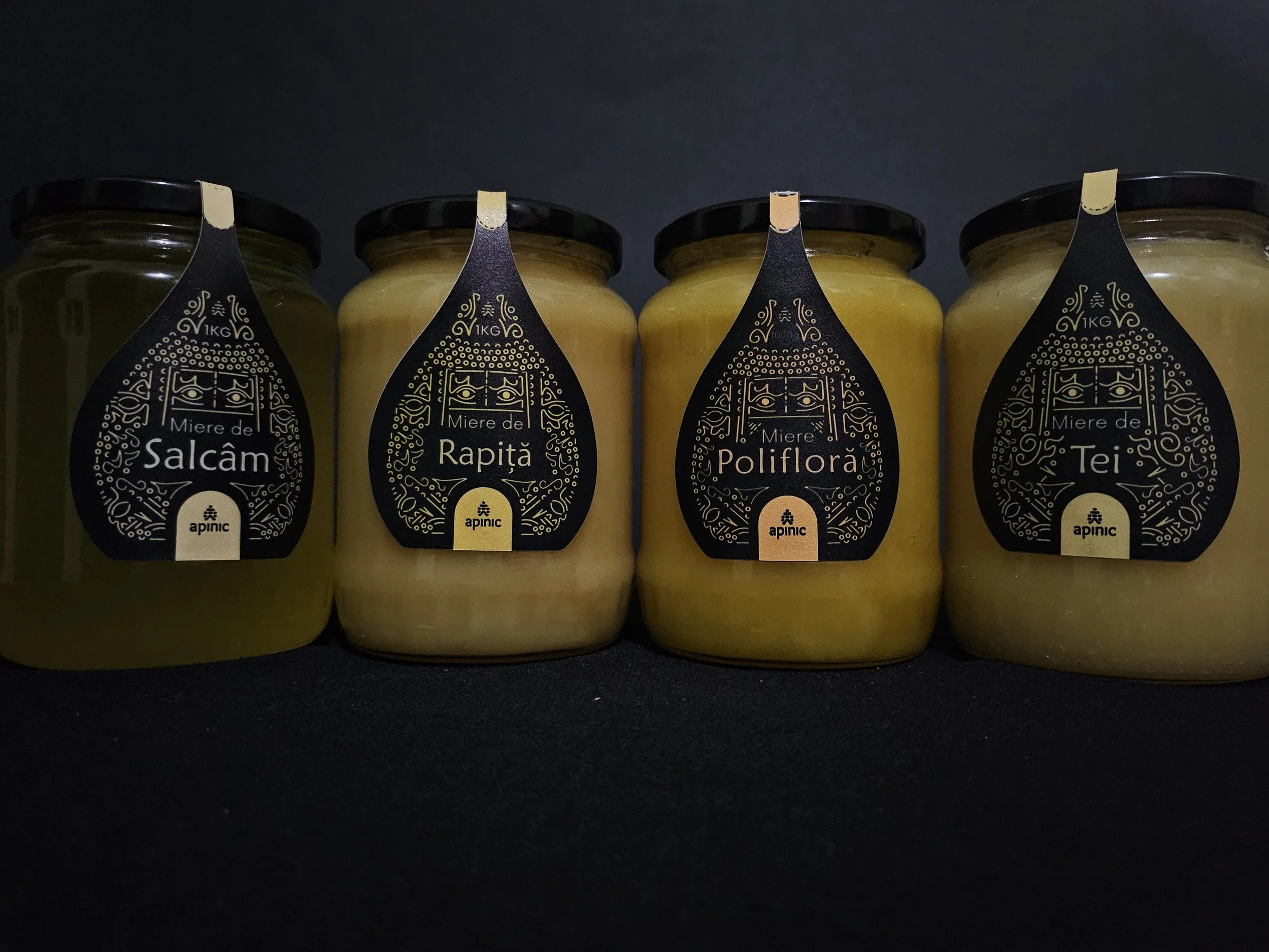 Four glass jars with black lids containing different types of honey, each with a black label featuring gold decorative design and text in Romanian.
