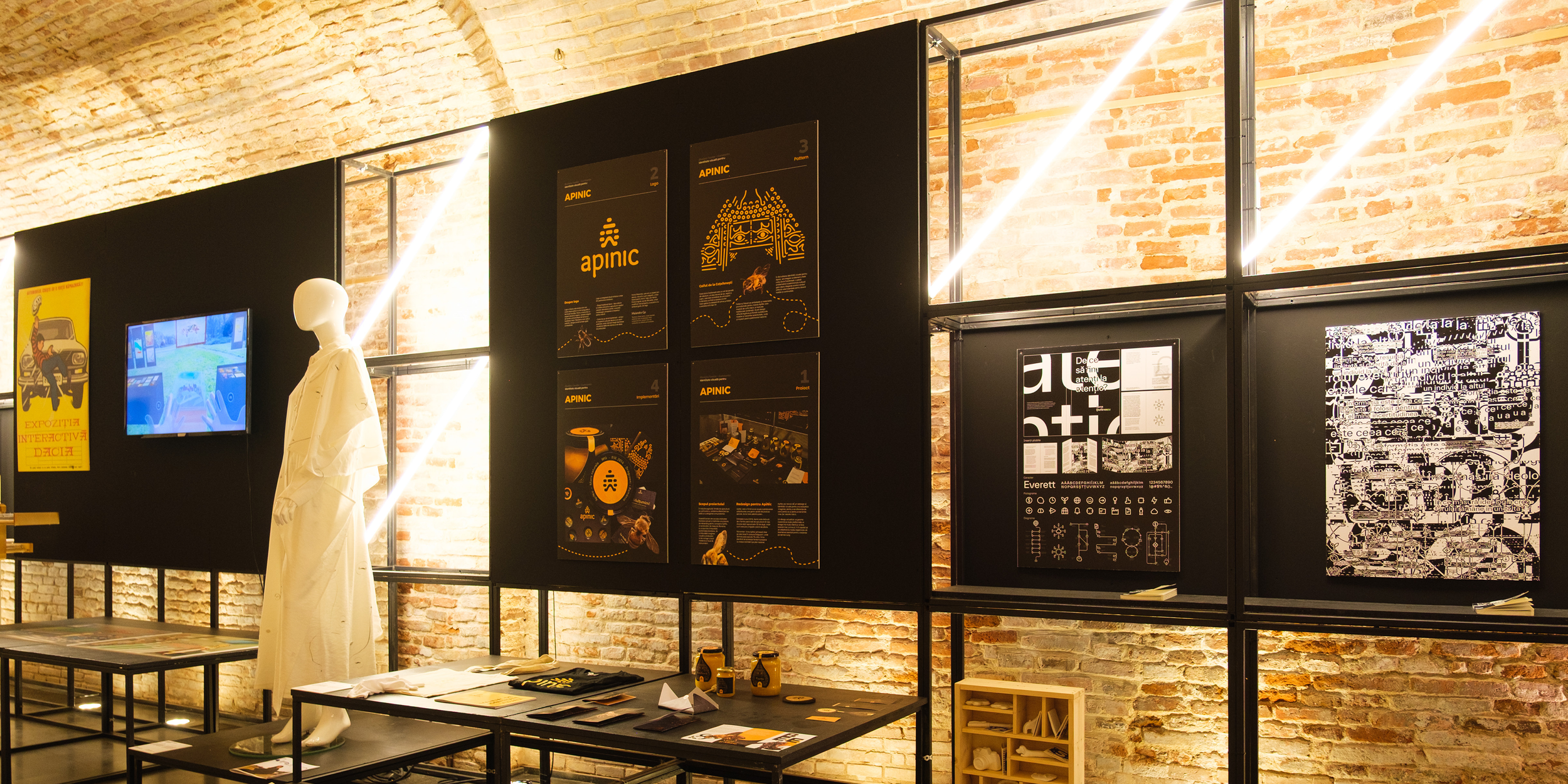 Exhibition space with a brick wall, black display panels featuring posters and artwork, a mannequin dressed in white, and a small table with pamphlets, in an art gallery setting.
