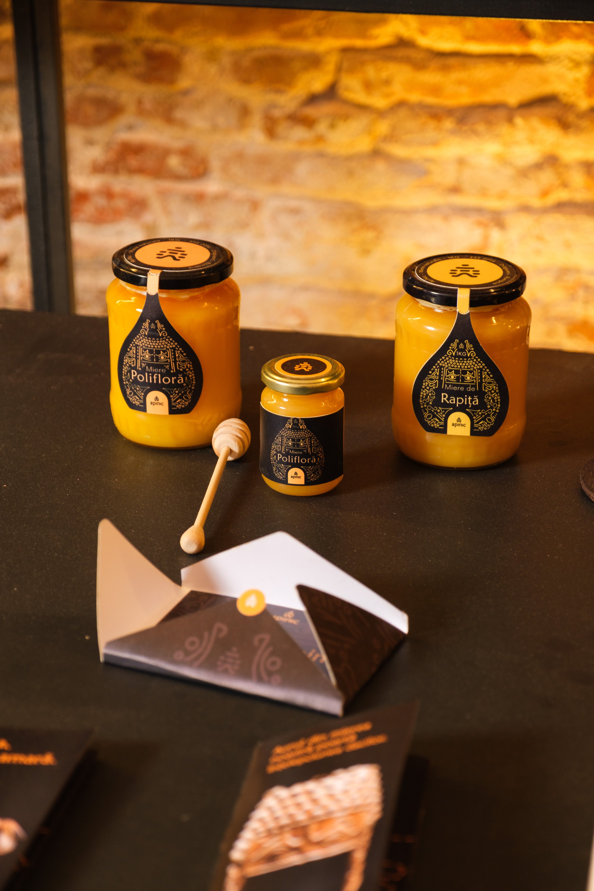 Three jars of honey labeled 'Poliflora', 'Rapti', and another smaller jar with black and gold labels and a honey dipper, set on a black surface against a brick wall background.