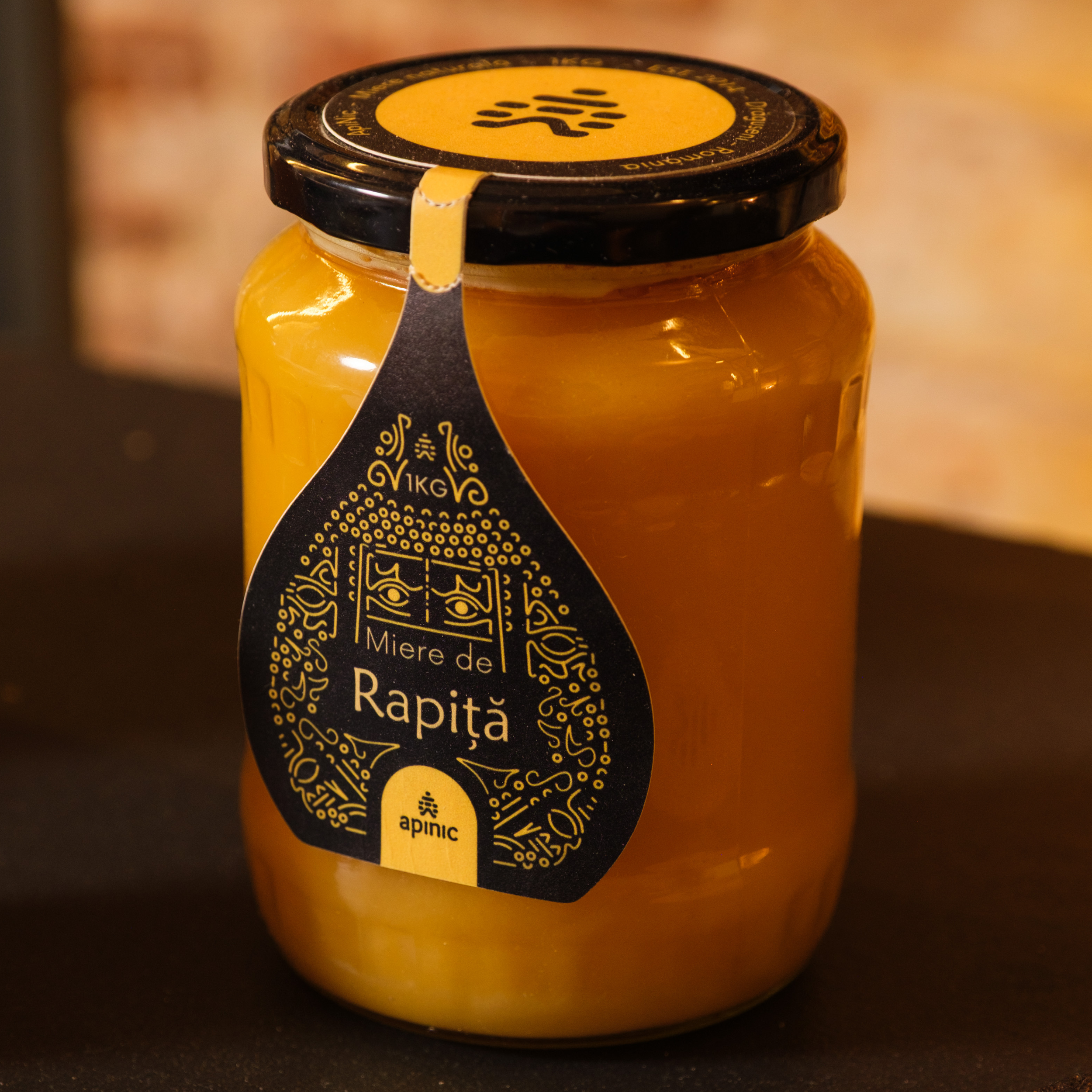 A glass jar with a black lid and a yellow label containing black decorative design and the words "Raptiä Mie de R" on it. The jar appears to contain a yellow substance, possibly honey or jam.