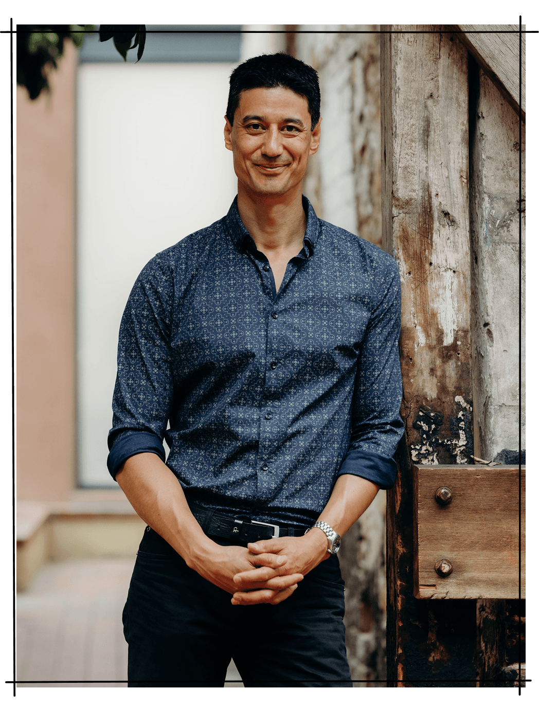 A portrait photo of Daniel Sih with his hands folded leaning on a tree.