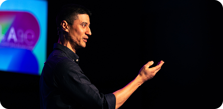 A person speaking or presenting on stage with a dark background, illuminated by stage lighting, with a blurred colorful screen behind them.