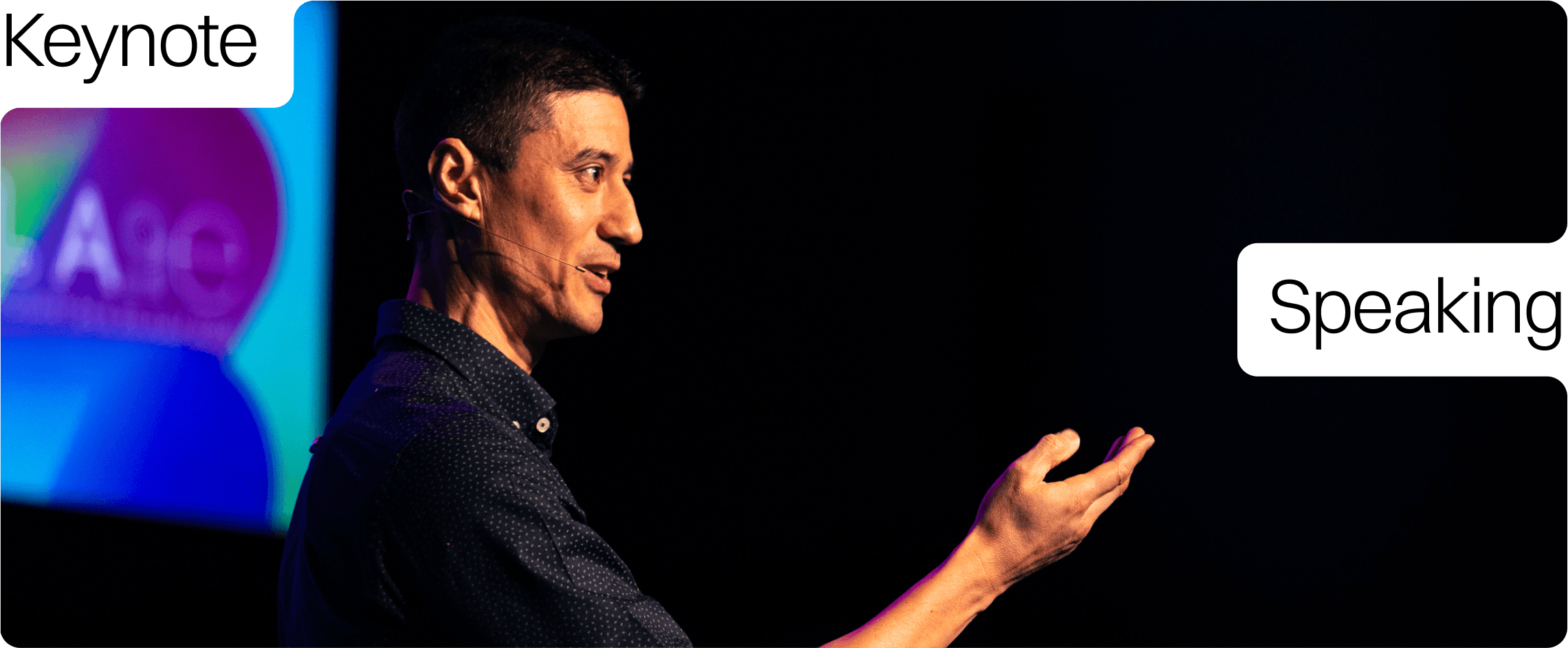 Daniel Sih in a dark dotted shirt standing on stage, speaking and gesturing with his hand during a presentation, with a colorful screen displaying 'AI' in the background.