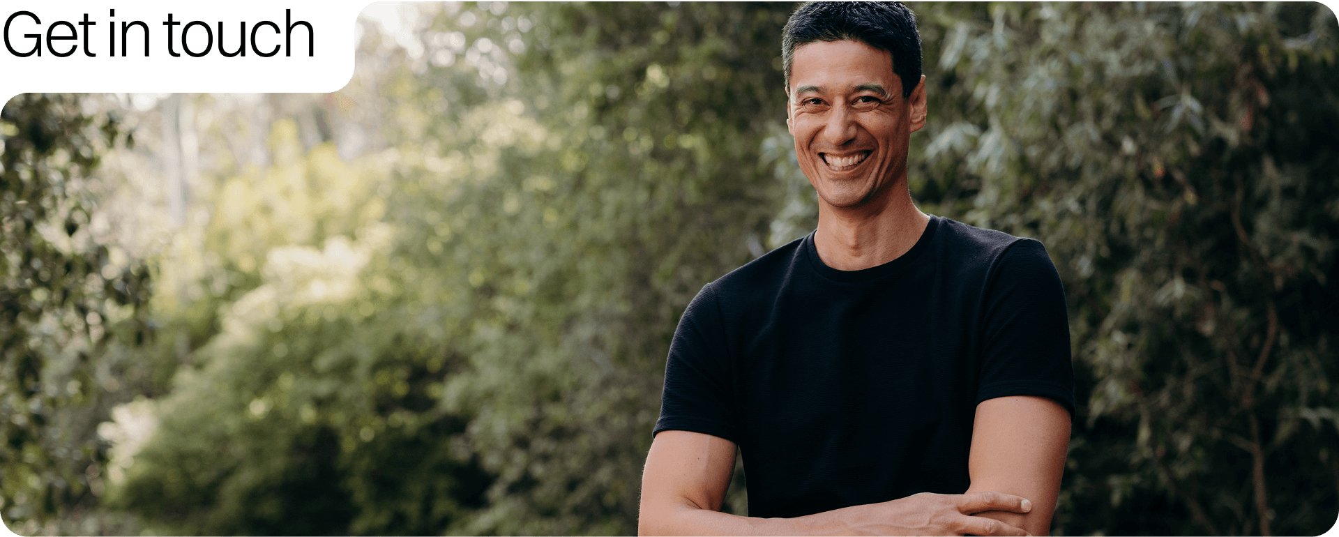 Daniel Sih smiling outdoors with arms crossed, wearing a black t-shirt, and surrounded by greenery.