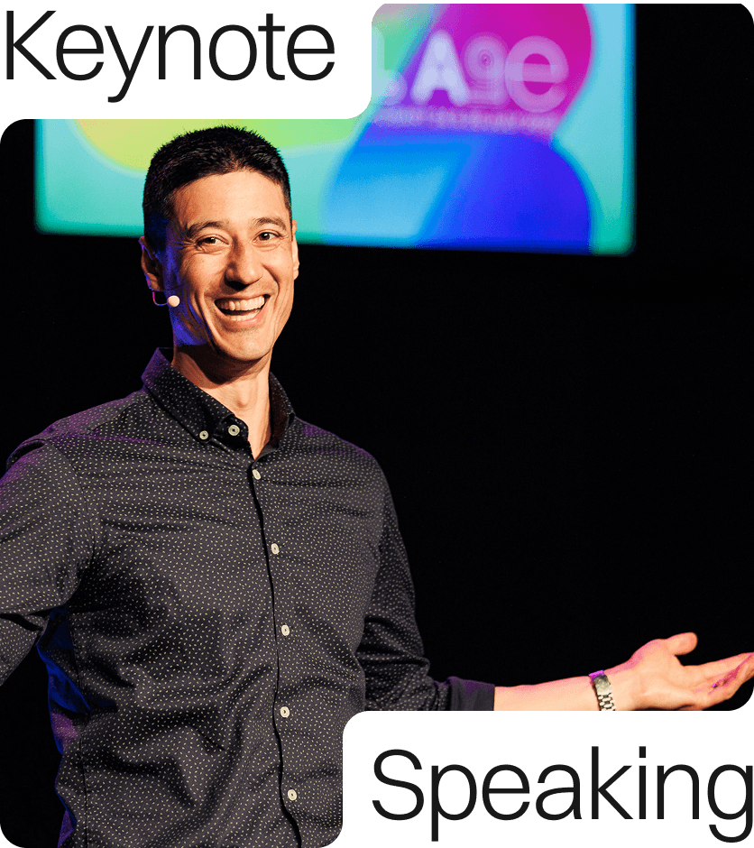 Daniel Sih wears a black speckled shirt while smiling and talking on stage. A brightly coloured screen is visible in the background.