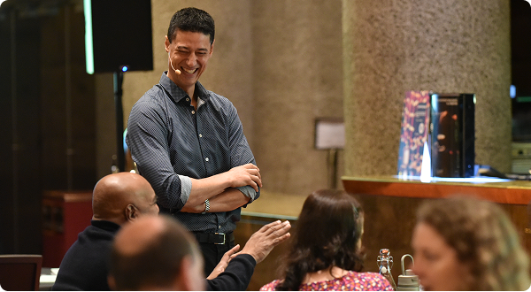 A man standing and smiling while speaking to a group of people seated at a table in a restaurant.