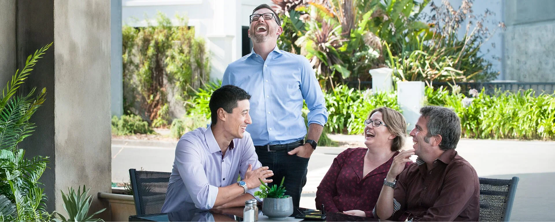 Four people are laughing and talking at an outdoor table, with greenery and plants in the background.