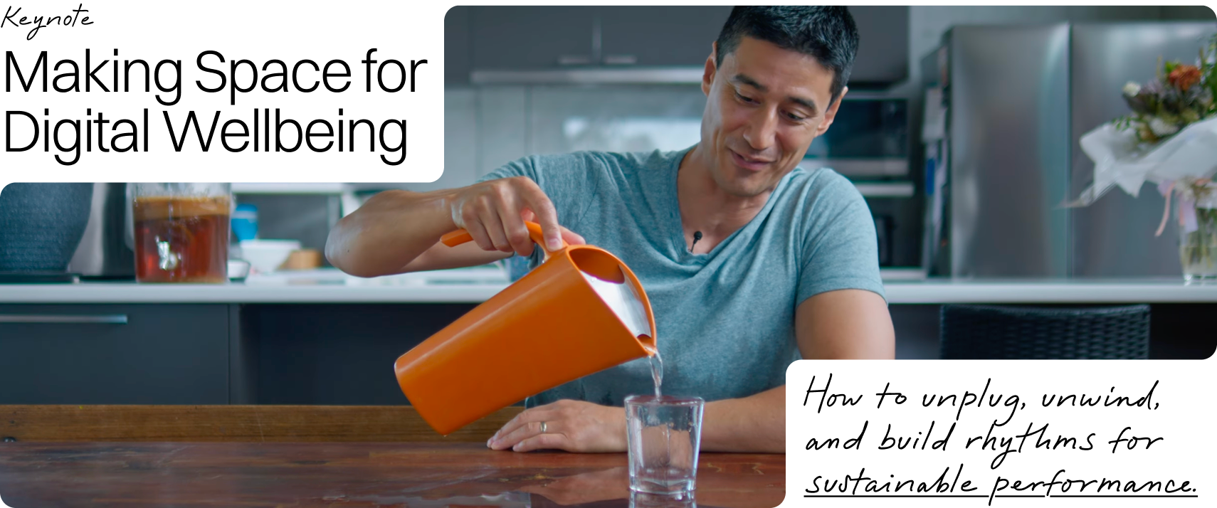 A man in a gray t-shirt pours water from an orange pitcher into a glass on a wooden table in a modern kitchen.