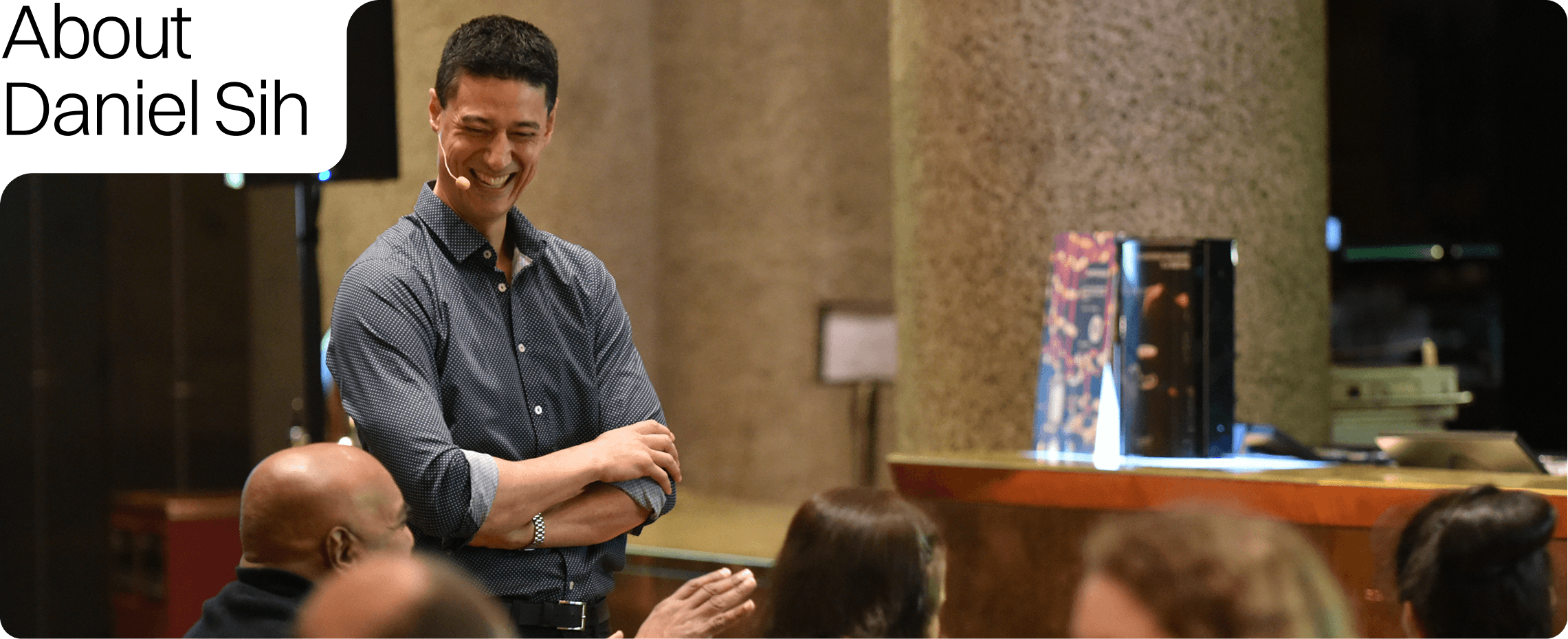 Text reads 'About Daniel Sih'. A man with short dark hair wearing a blue patterned shirt smiling and laughing in a conference or seminar setting, with people seated around him and a person in the foreground with a shaved head.