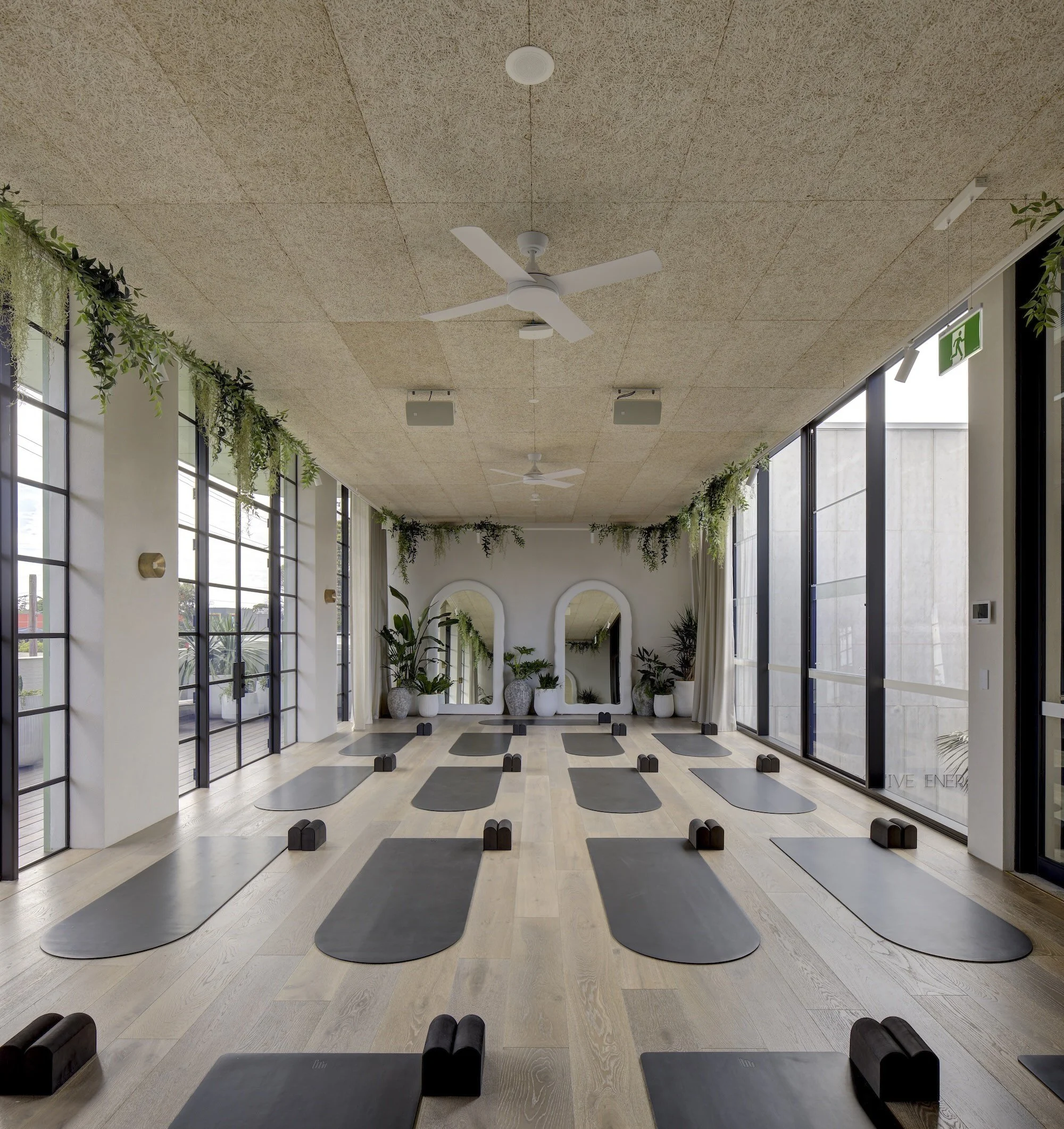 Indoor yoga or meditation studio with mats and small blocks arranged on a wooden floor, large windows with curtains and greenery, ceiling fans, and wall-mounted speakers.