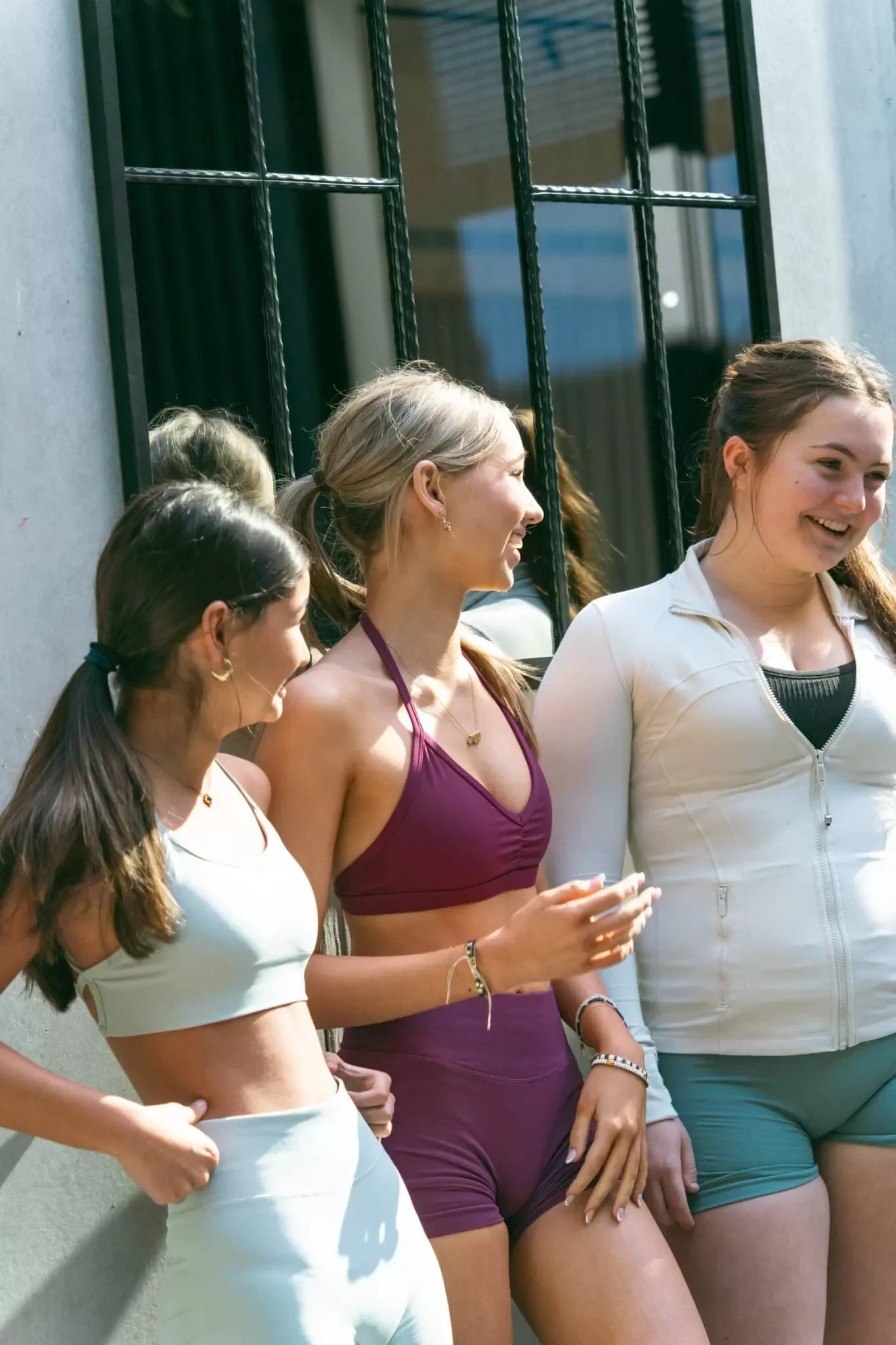 A group of women standing close together outside, smiling and talking, dressed in casual athletic wear, with a building with black window frames in the background.