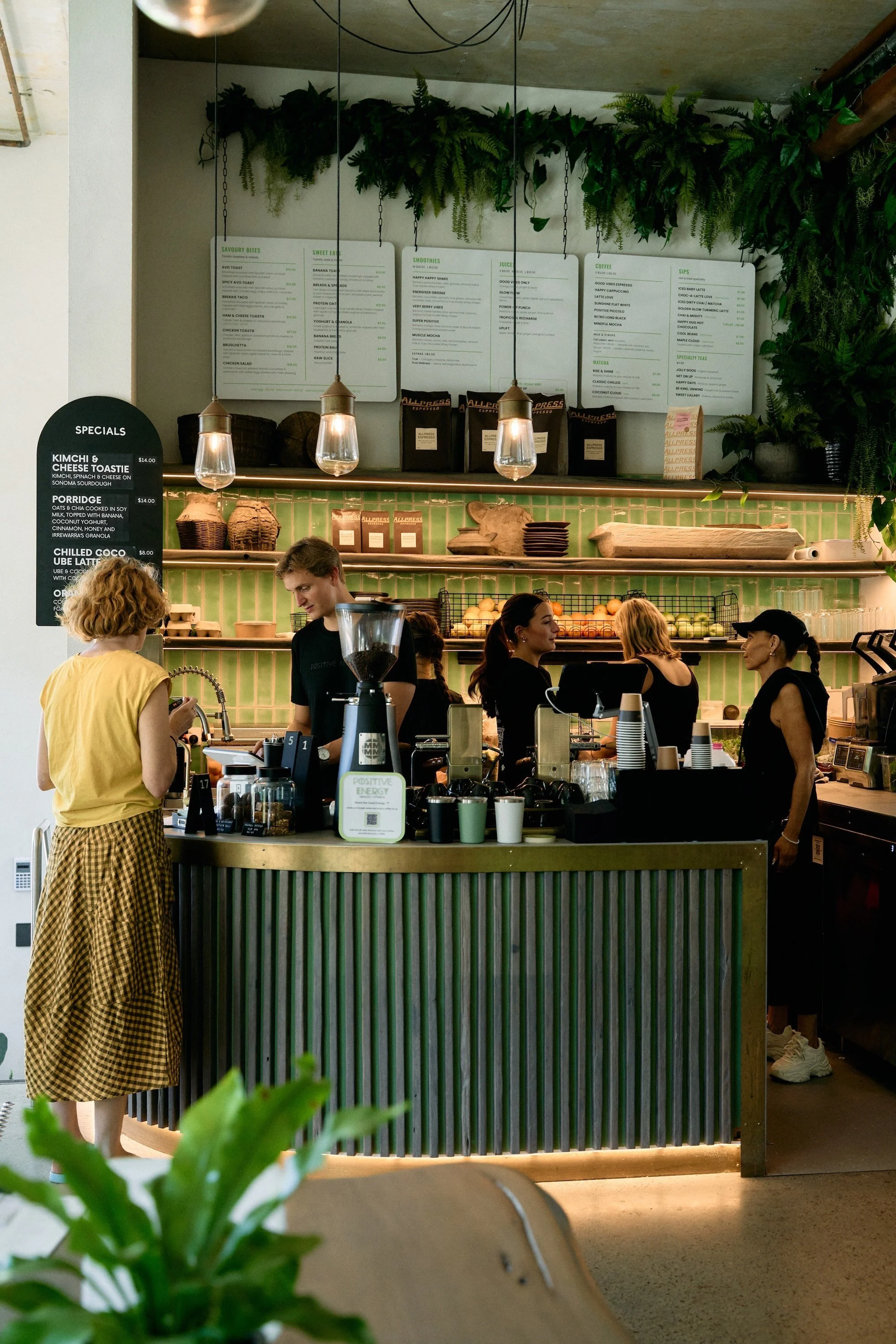 Interior of a coffee shop counter with three people working behind it, green tiled wall, hanging pendant lights, menu boards above, and lush green plants on top of the counter.