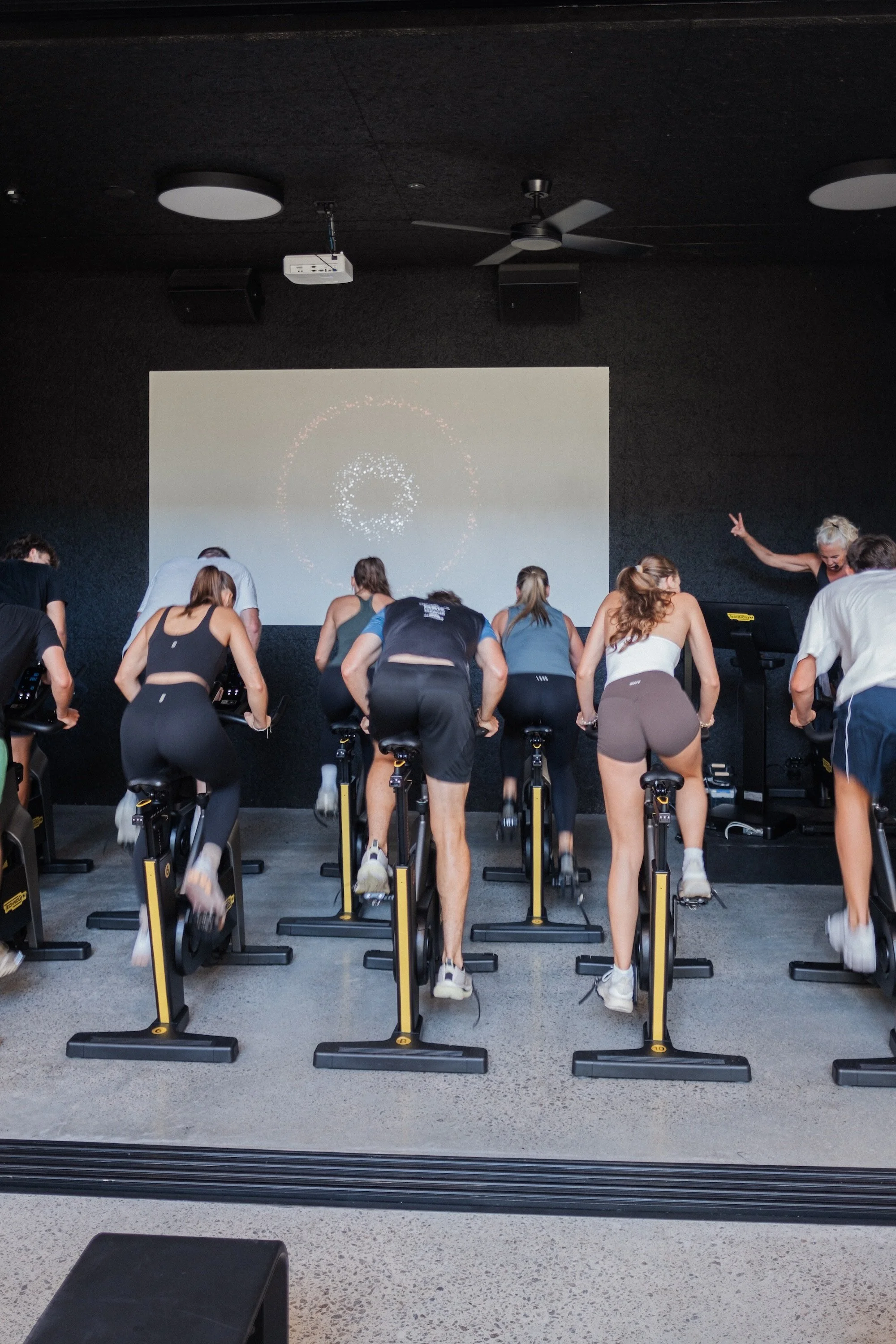 People participating in a spin class on exercise bikes in a dark room with a large screen on the front wall showing a circular design, instructor leading the class, some participants raising their hands.