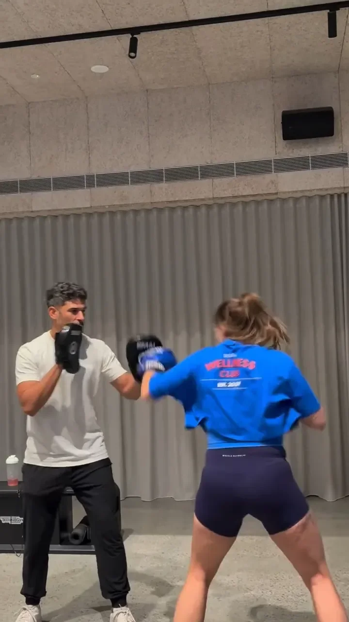 A woman practicing boxing with a trainer in an indoor gym, punching focus mitts held by the trainer.