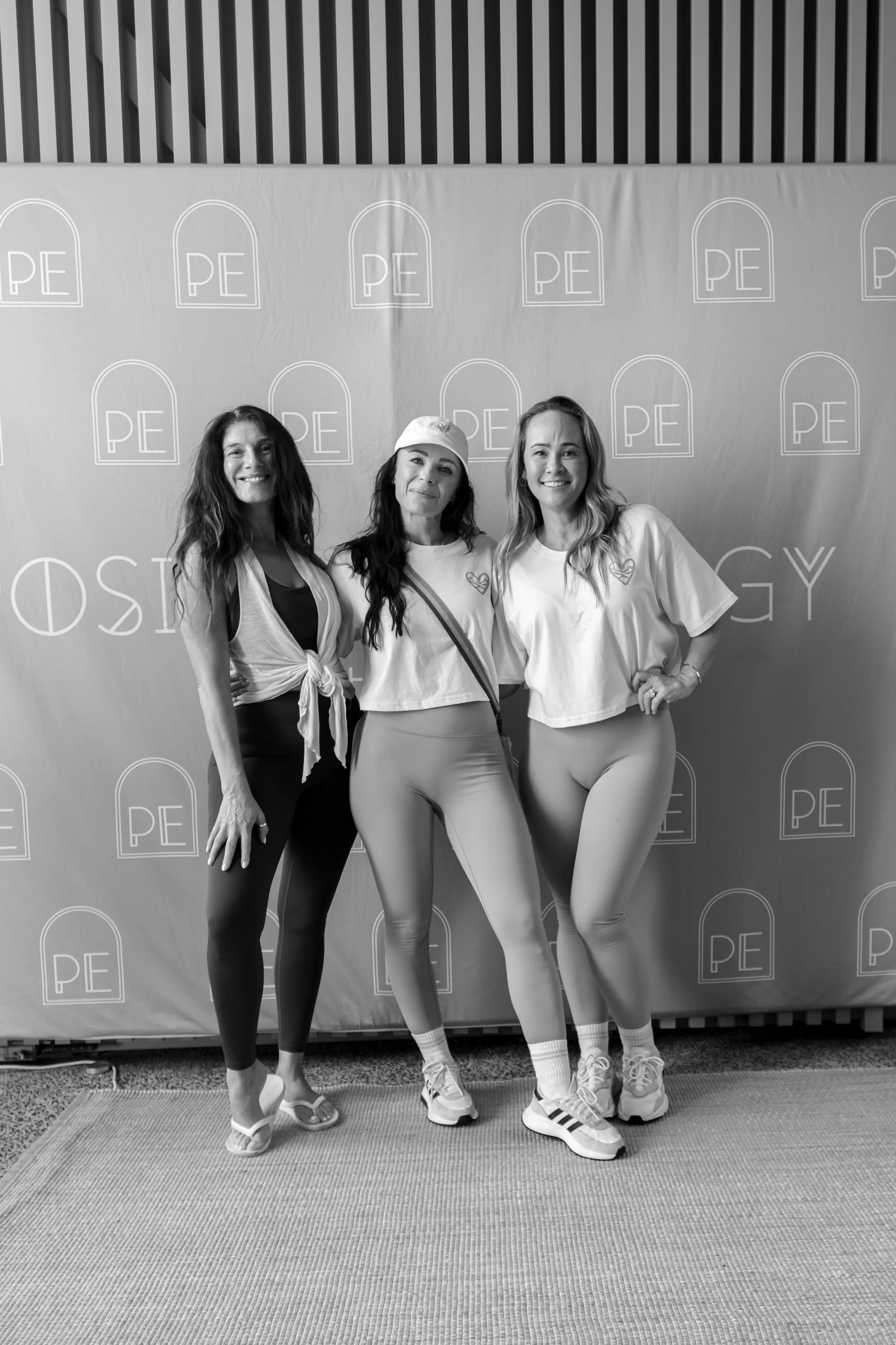 Three women standing in front of a backdrop with repeated PE logos, smiling and posing for the photo.