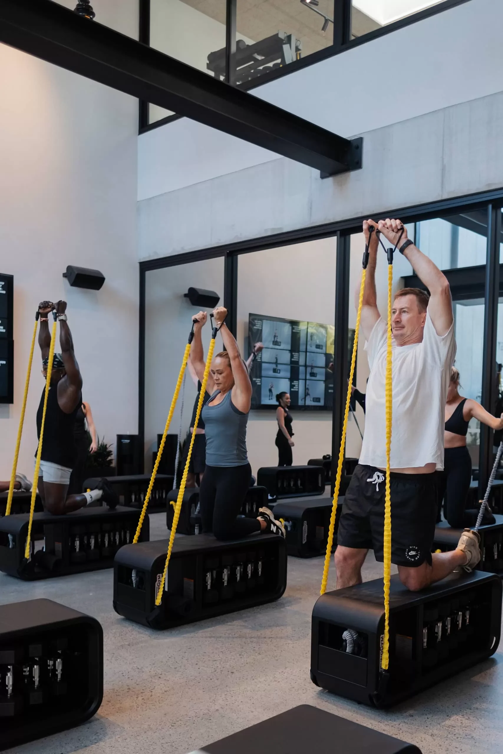 People participating in a fitness class using TRX suspension trainers, kneeling on workout benches in a modern gym with mirrors and wall-mounted screens.