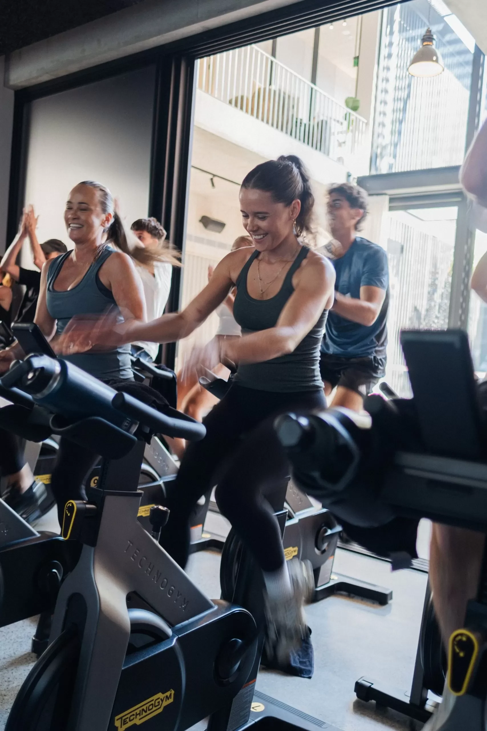 People exercising on stationary bikes in a modern gym with large windows and natural light.