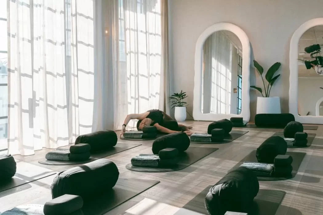 Yoga studio with mats, bolsters, and blankets set up for a class, sunlight coming through large windows, mirrors on the wall, and a person in a yoga pose.