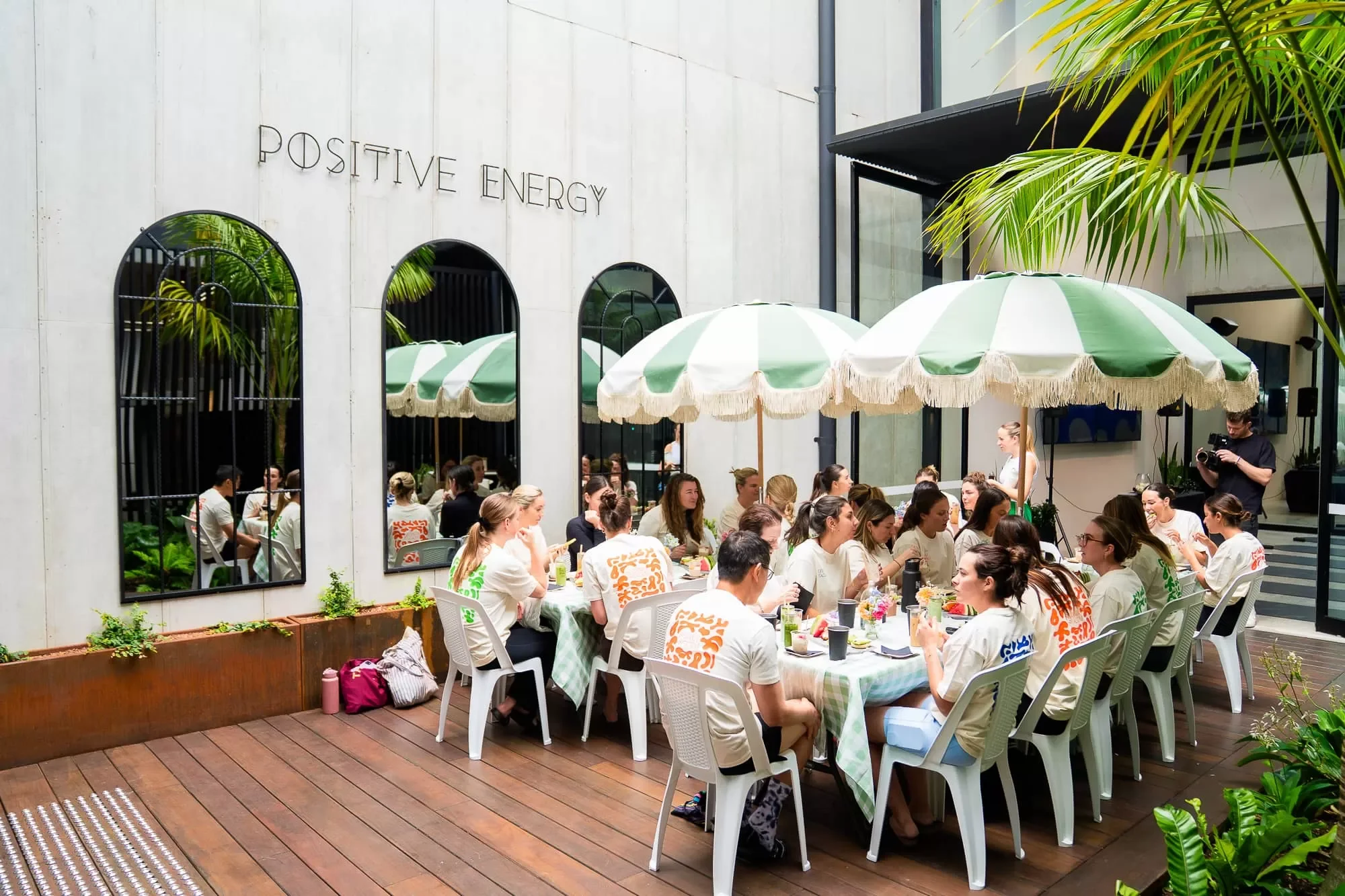 People sitting at a long outdoor table with green and white checkered tablecloths, under a green and white striped umbrella, in a modern courtyard with palm trees and large windows, with a sign that reads positive energy.