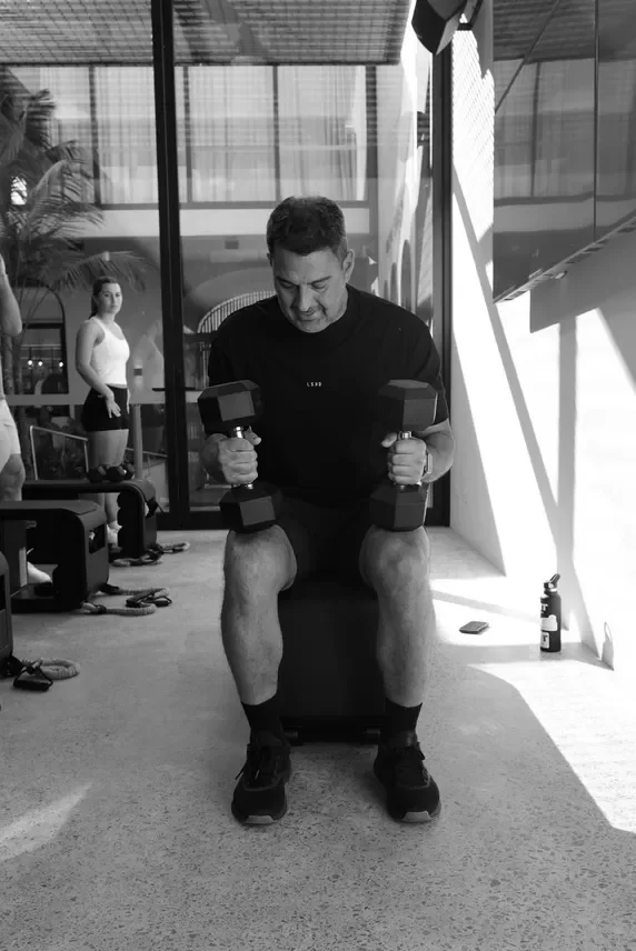 A man sitting on a bench lifting dumbbells in each hand, with a woman standing in the background in a gym or fitness studio.