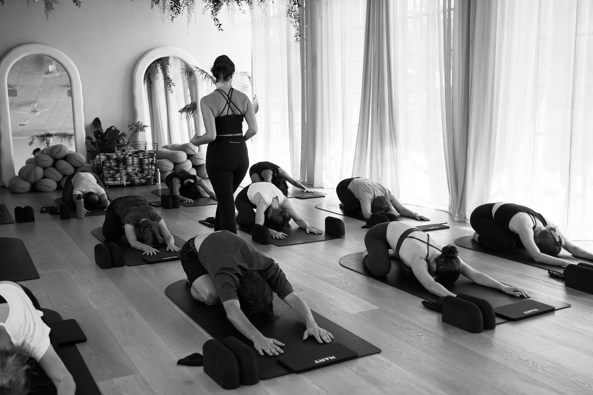 A group of people practicing yoga in a studio. They are in a forward bend pose on yoga mats, following an instructor who stands at the front of the room. The room has large windows with curtains and mirrors on the wall, and some stacked pillows and plants are visible.