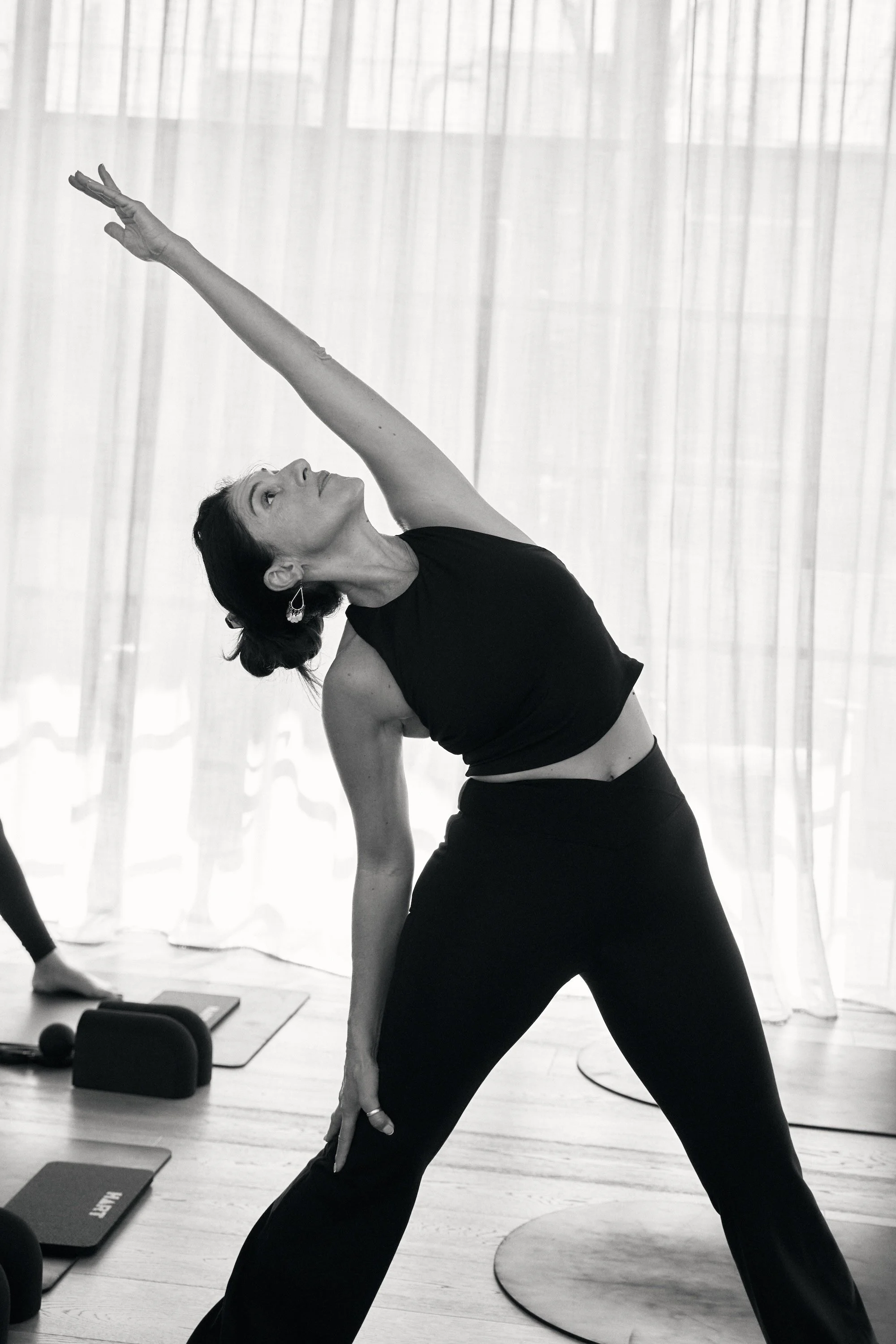 A woman practicing yoga indoors, performing a side stretch pose, with exercise mats, weights, and a large window with curtains in the background.