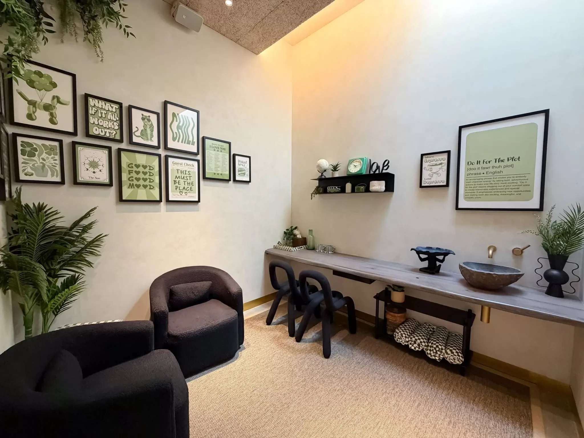 A cozy room with a beige carpet and white walls decorated with framed wall art and plants. There are two black armchairs and a stool around a wooden table, with a shelf holding decorative items and a large framed quote on the wall.