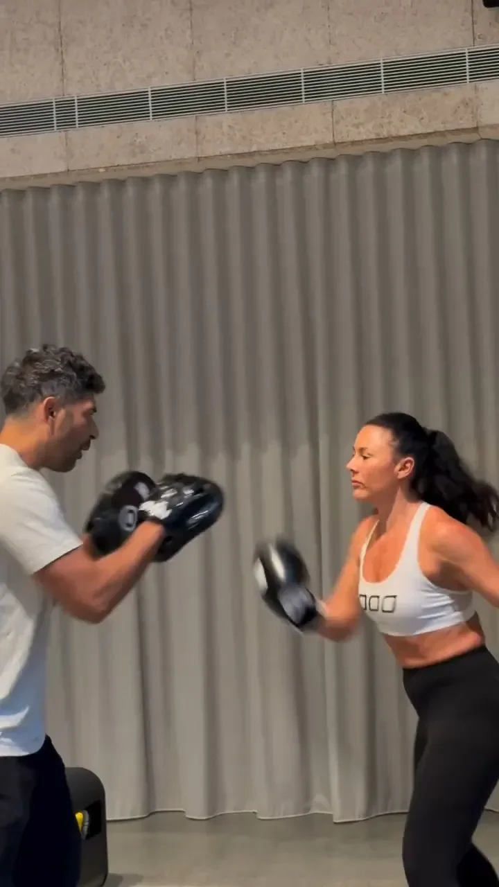 A woman is boxing with a man holding focus mitts indoors in front of a gray curtain.