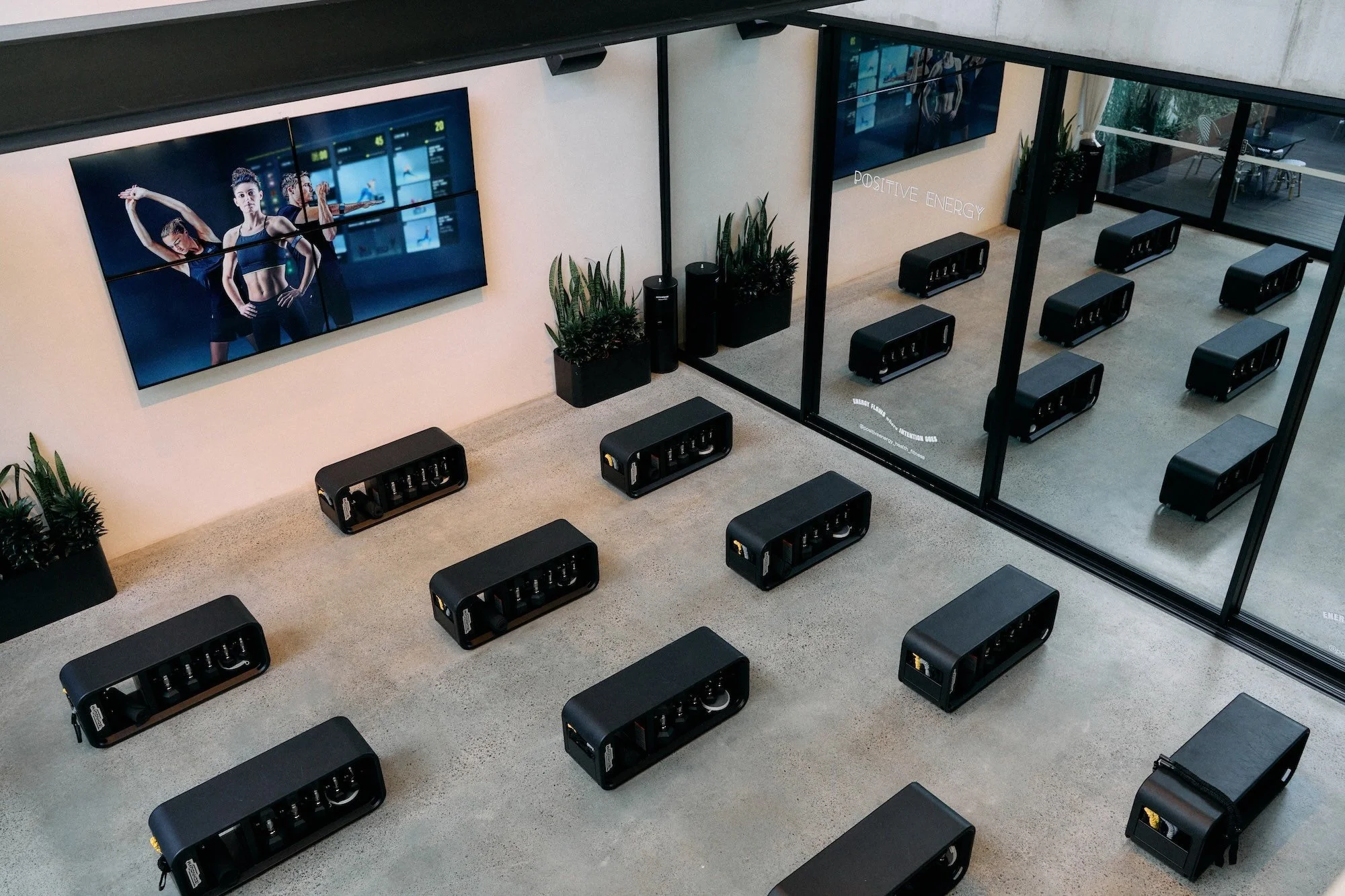 Interior view of a fitness or wellness space with multiple black massage chairs arranged in a grid pattern, large flat screen monitors on the walls displaying fitness or dance videos, plants, and glass walls with the words 'Positive Energy' on the glass.