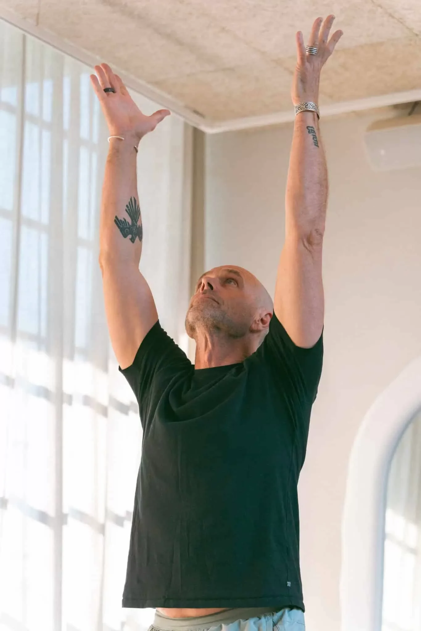 A man with tattoos on his arms, wearing a black shirt, is raising his arms above his head indoors, with his eyes looking up. The background shows tall windows with sheer white curtains.