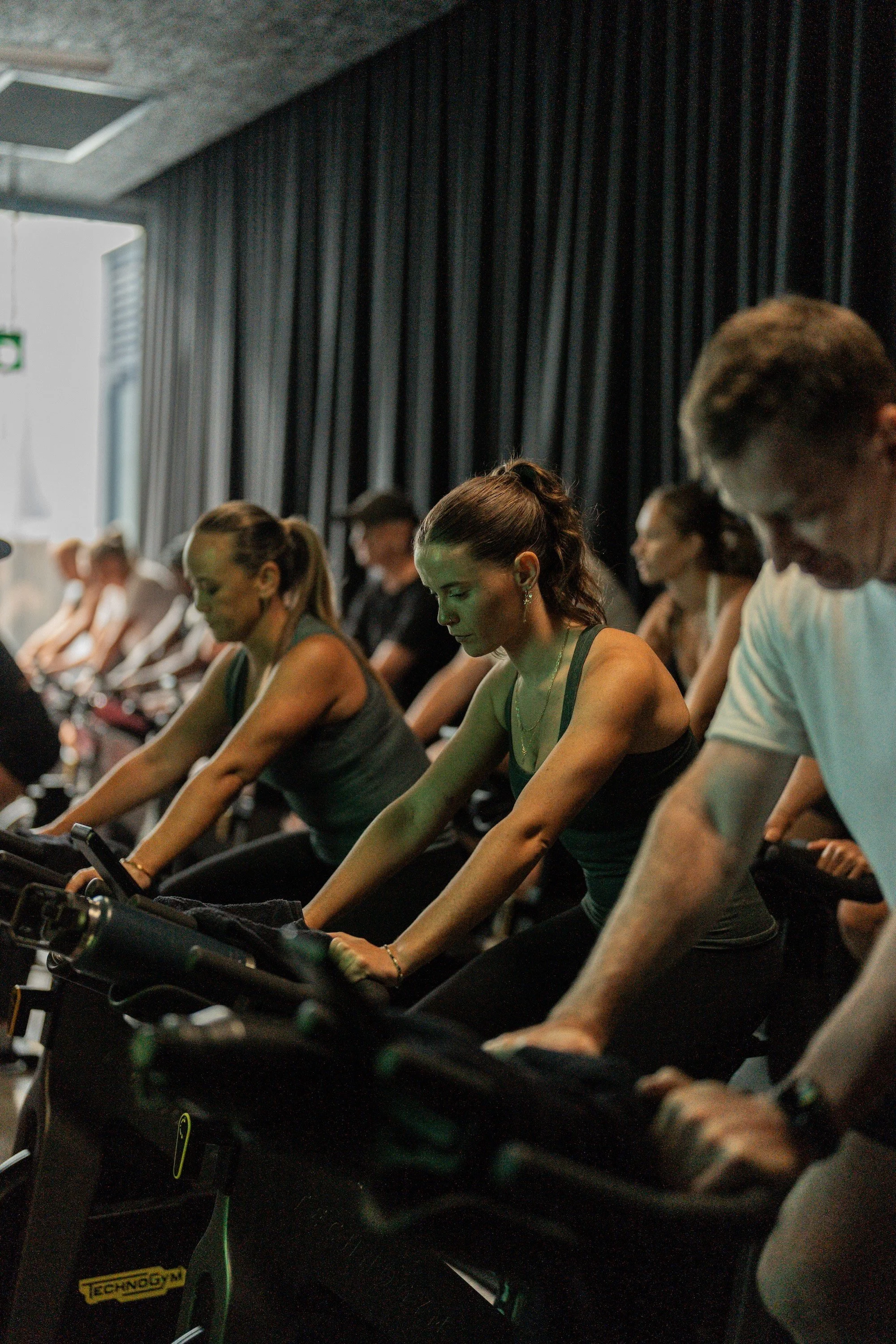 People participating in a spinning class, riding stationary bikes in a dimly lit room with dark curtains.