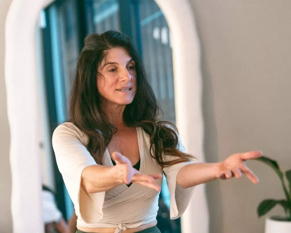 A woman with long dark hair and light skin, gesturing with her hands while speaking indoors.