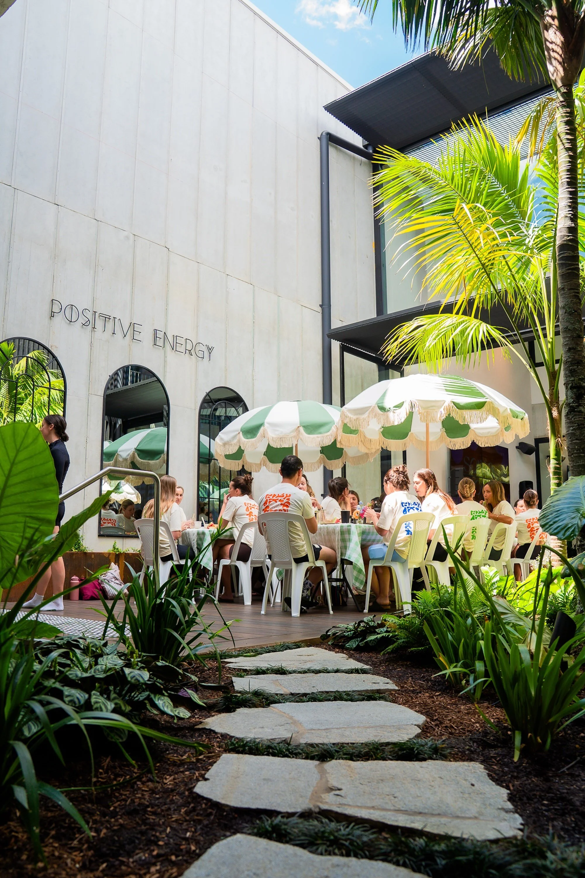 Outdoor patio scene with people sitting at tables under umbrellas, surrounded by lush green plants and trees. Building with large windows and the words 'Positive Energy' on the wall visible in the background.