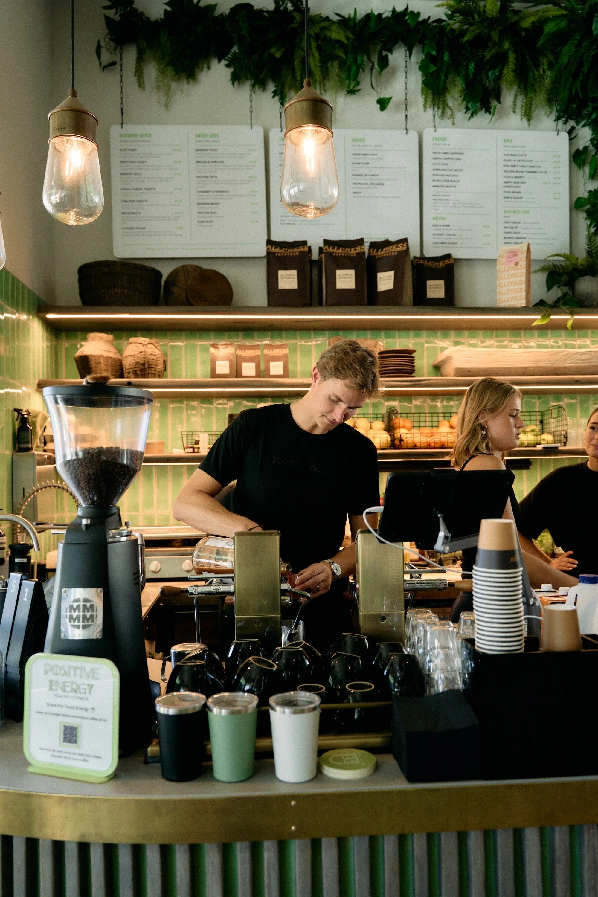 View of a coffee shop counter with baristas preparing drinks, green tiled wall, shelves with baskets, and a menu board in the background.