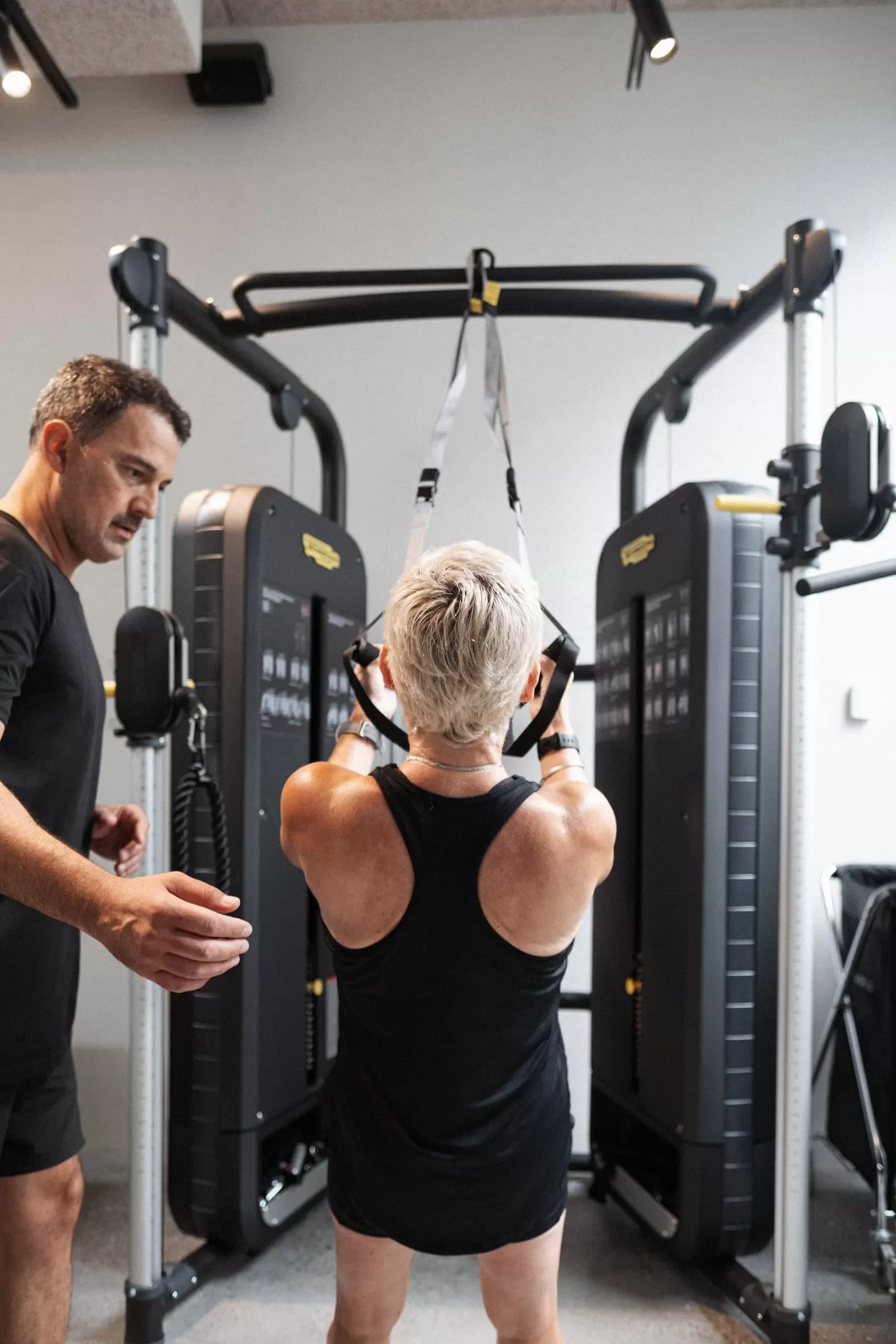 A woman lifting weights in a gym, assisted by a man standing nearby. The gym equipment includes a pull-up bar and a cable machine.