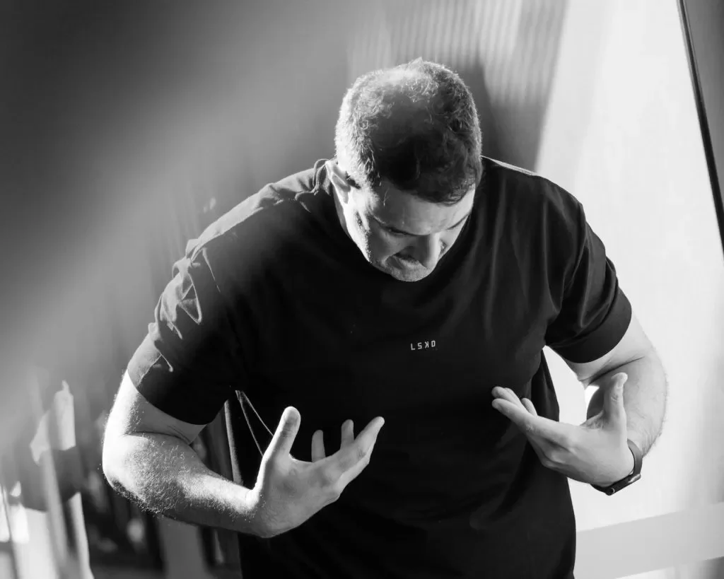 A man in a black t-shirt with his head bowed and hands pointing to his chest, standing indoors with light coming from the right side.