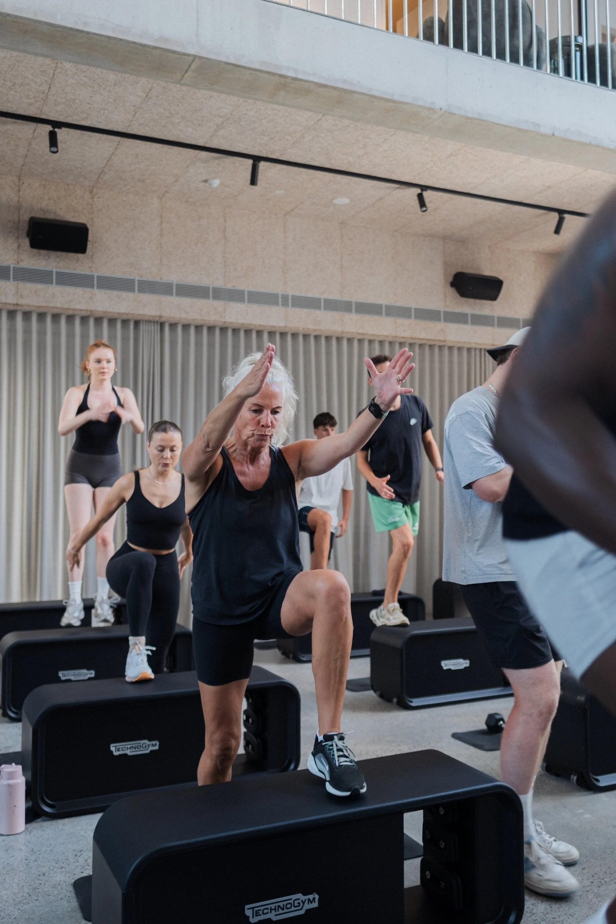 People participating in a step aerobics class in a gym, stepping on black step platforms, with a woman leading the class in the foreground.