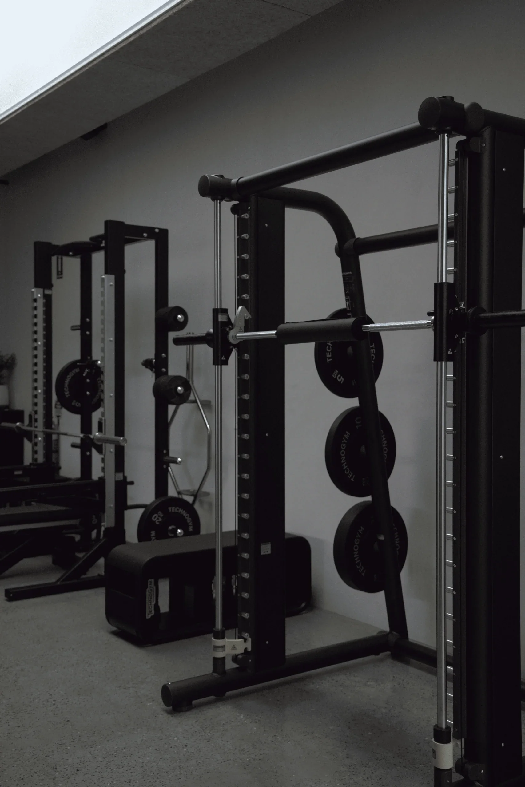 A gym with weightlifting equipment, including a squat rack with weight plates, and a black padded bench on a gray carpeted floor.
