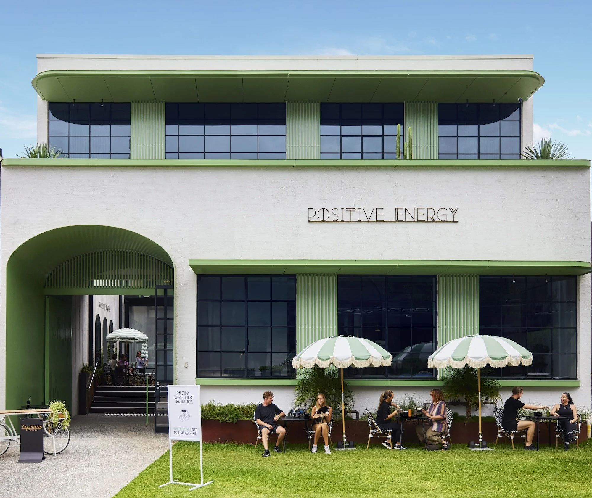 A modern building with the words 'Positive Energy' on the facade, featuring green accents and large glass windows. There is an outdoor seating area with people enjoying drinks under umbrellas, and a sign advertising smoothies, coffee juices, and healthy food.