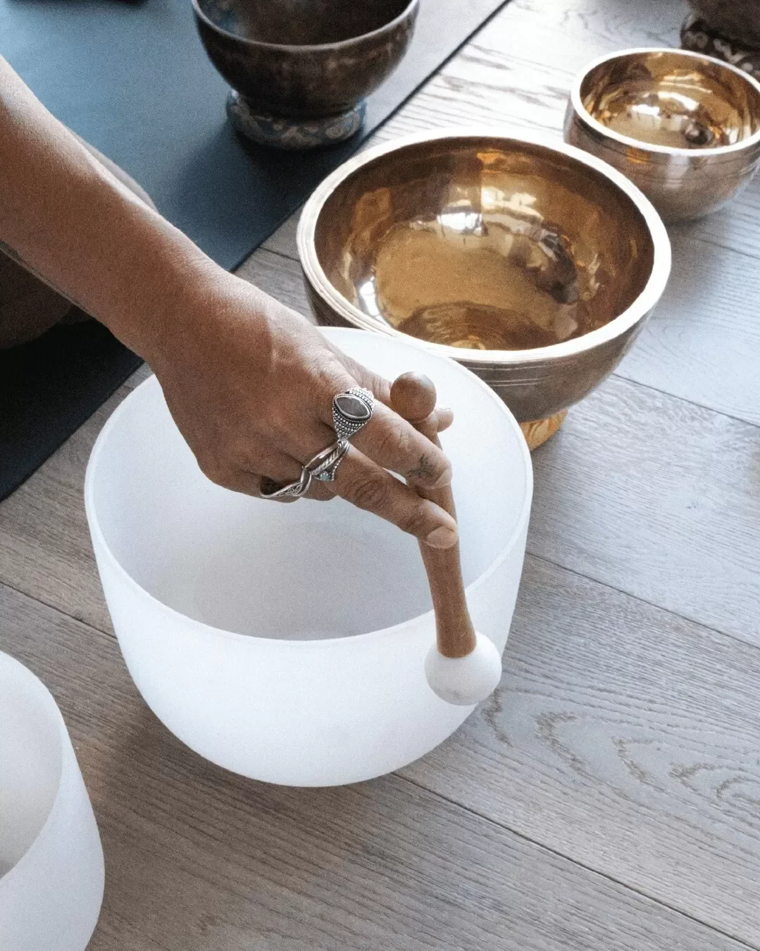 Person striking a balanced, seated meditation pose with fingers touching on a large, glowing white crystal singing bowl, surrounded by other metallic bowls on a wooden floor.