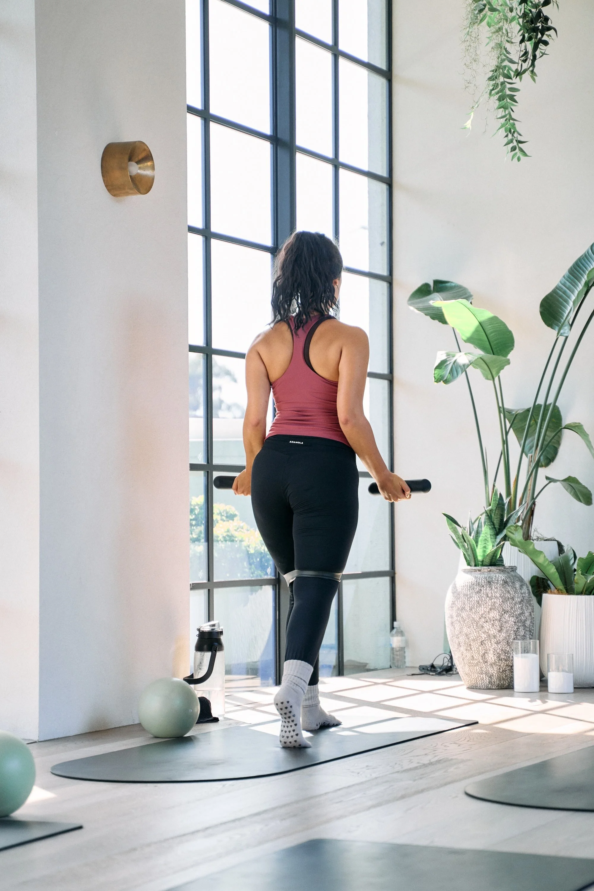 A woman in workout clothes holding a barbell in a bright, modern fitness studio with large windows and indoor plants.