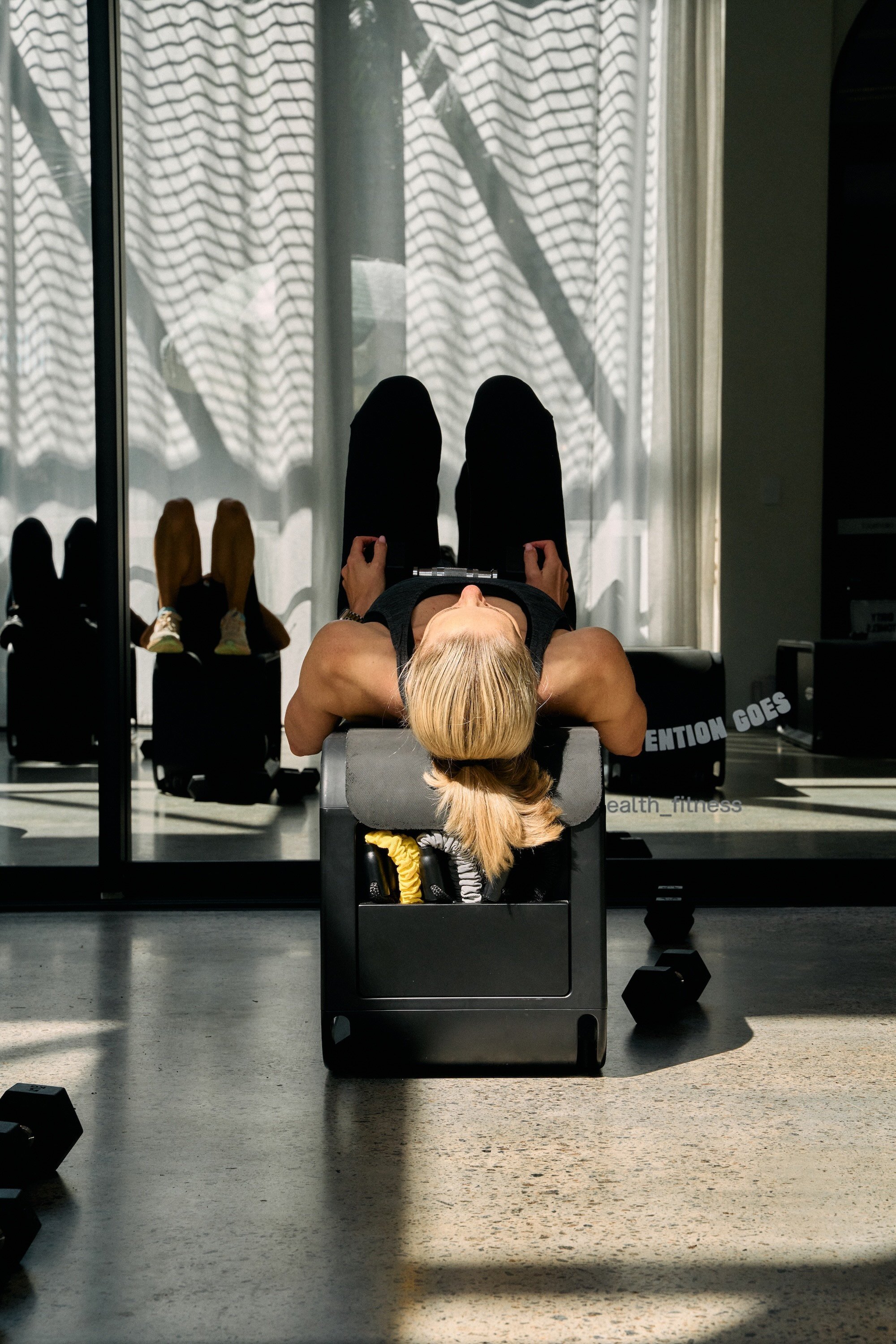 A woman lying on her back on a workout bench, holding a dumbbell in each hand, with her legs folded and raised in the air in a gym. There are shoes on a shelf behind her and a mirror reflecting her image. Sunlight is coming through the curtains, casting shadows on the floor.