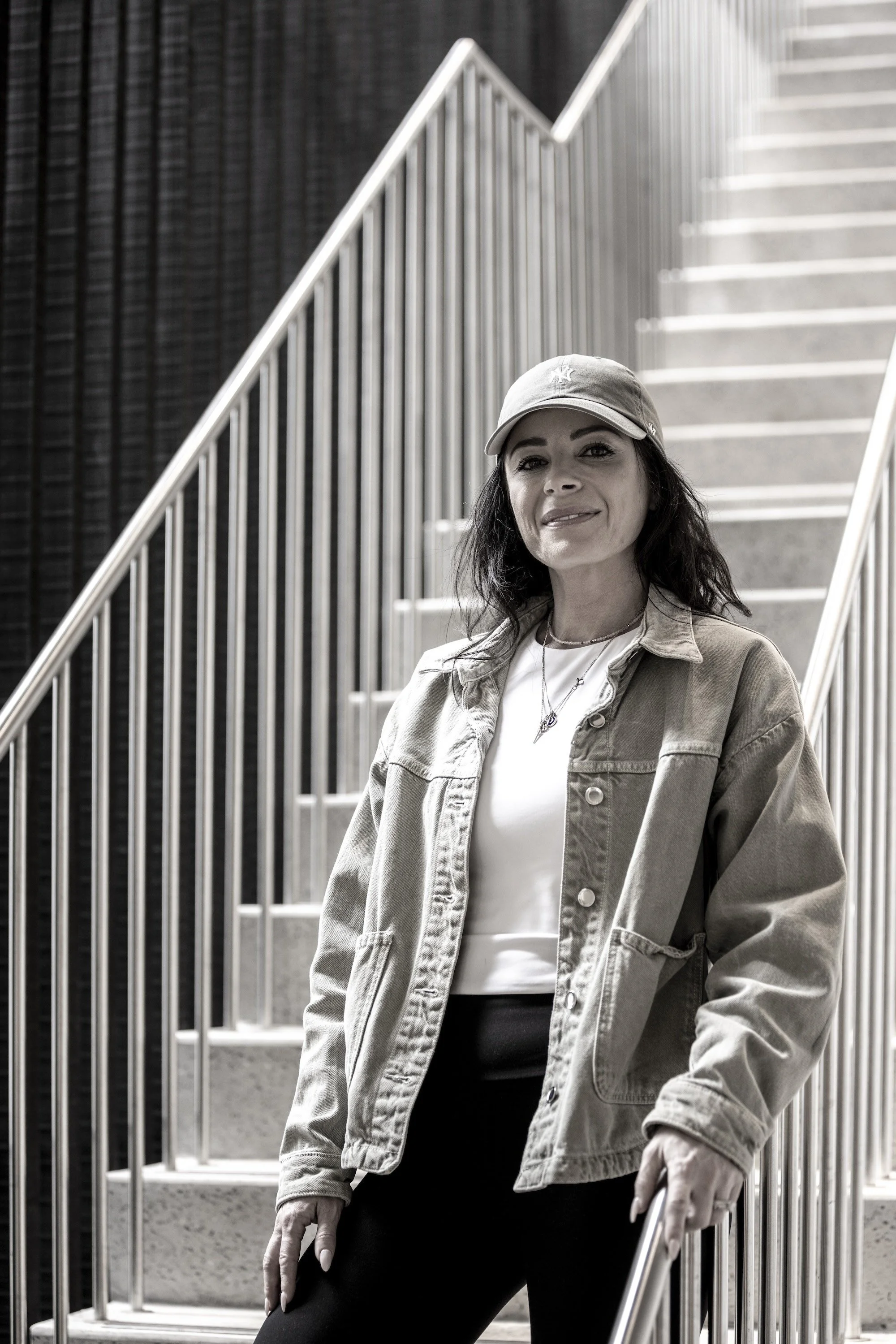 A woman wearing a baseball cap, white t-shirt, and denim jacket standing on a staircase with metal railing, smiling at the camera in black-and-white photo.