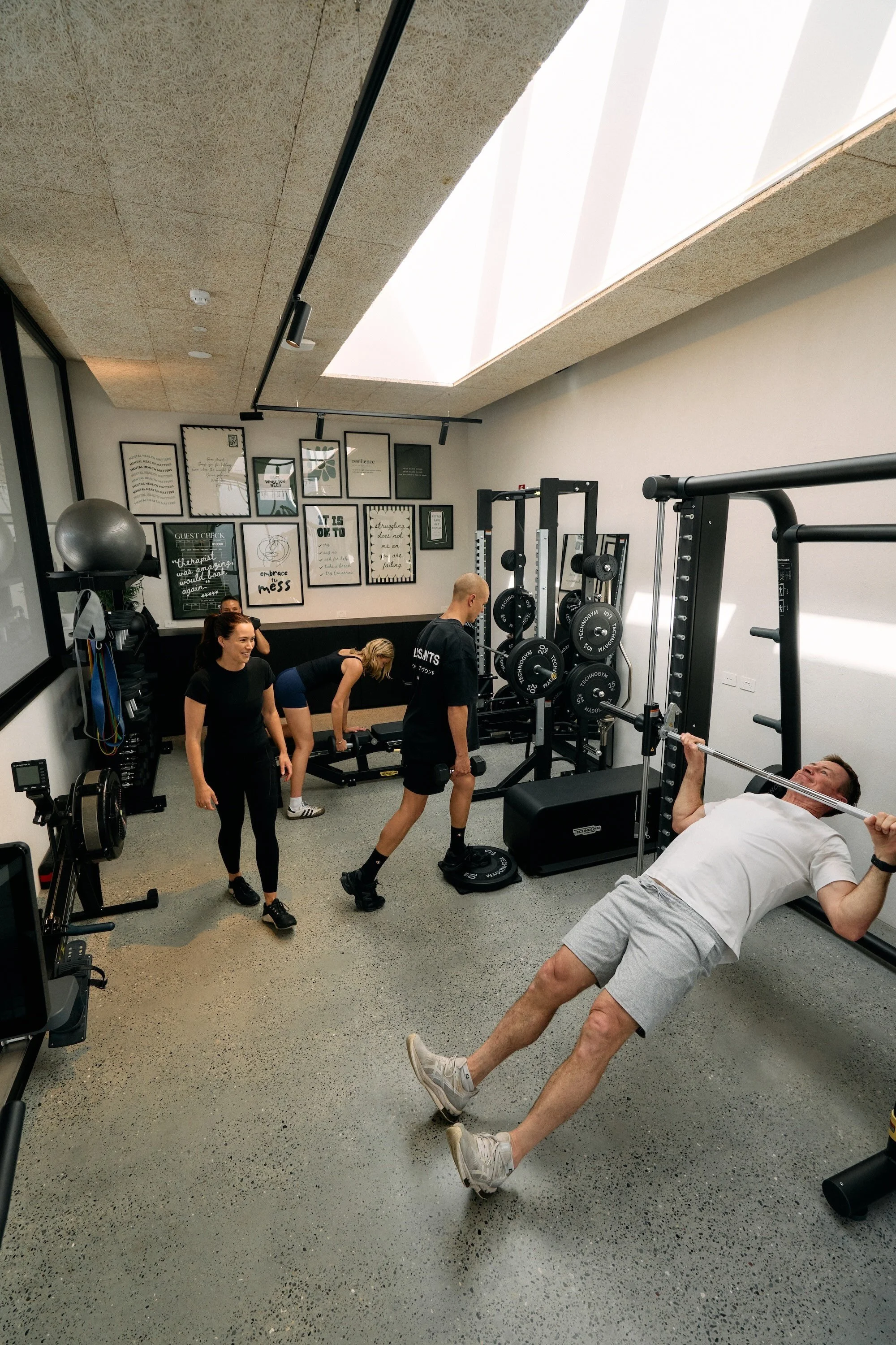 People working out in a gym, with some doing weightlifting, stretching, and others observing, inside a room with framed posters on the wall and a ceiling skylight.