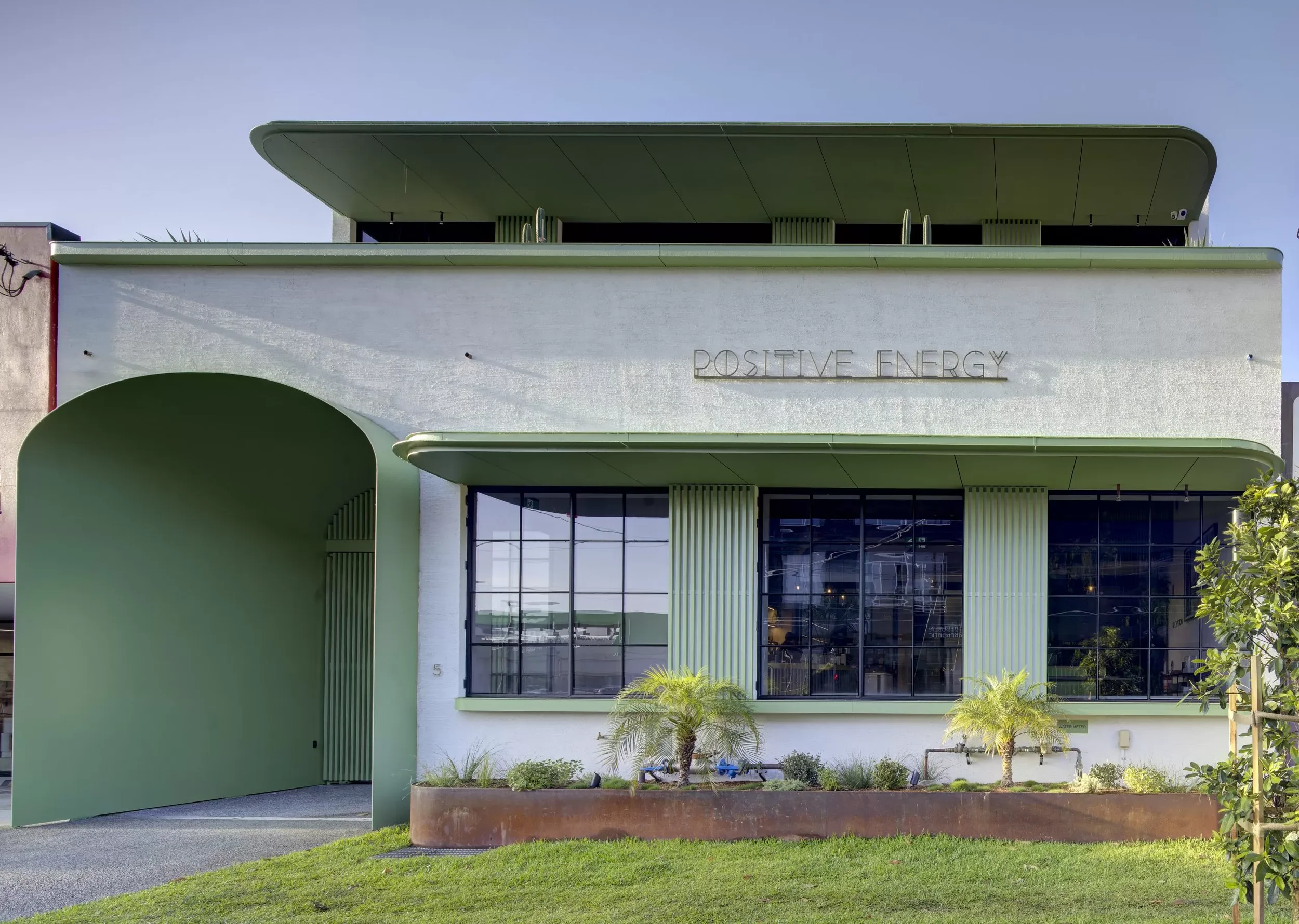 Modern building with green accents, large windows, and a sign that reads 'Positive Energy' on the front wall. Small trees and plants in front of the building, under a clear blue sky.