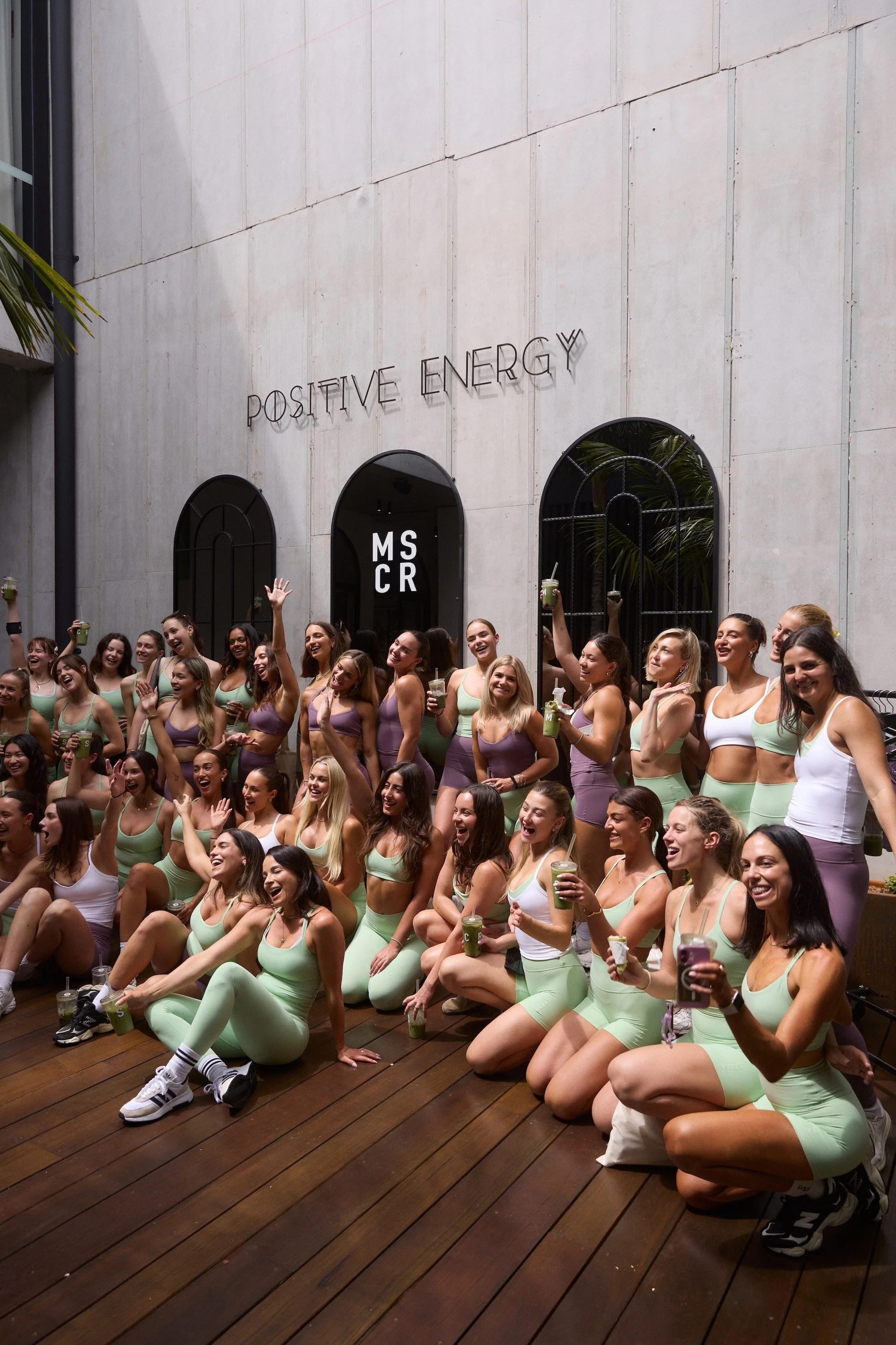 Group of smiling women in workout outfits sitting and kneeling on wooden floor at fitness event, holding drinks, in front of a plain wall with a sign that reads 'POSITIVE ENERGY' and arched windows