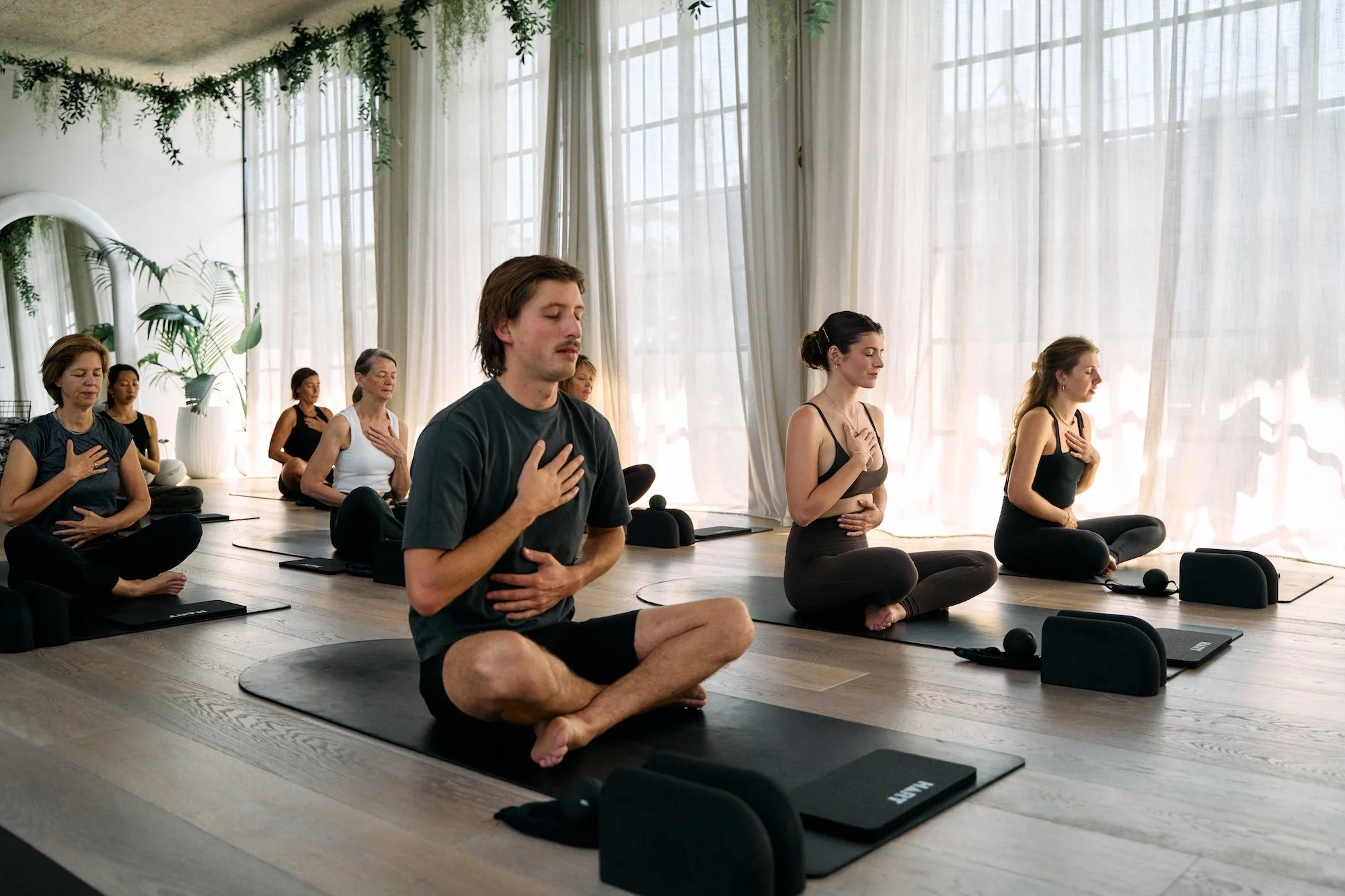 A group of people practicing meditation in a bright yoga studio with large windows and flowing curtains. They are seated cross-legged on mats, with hands on their chests and eyes closed.