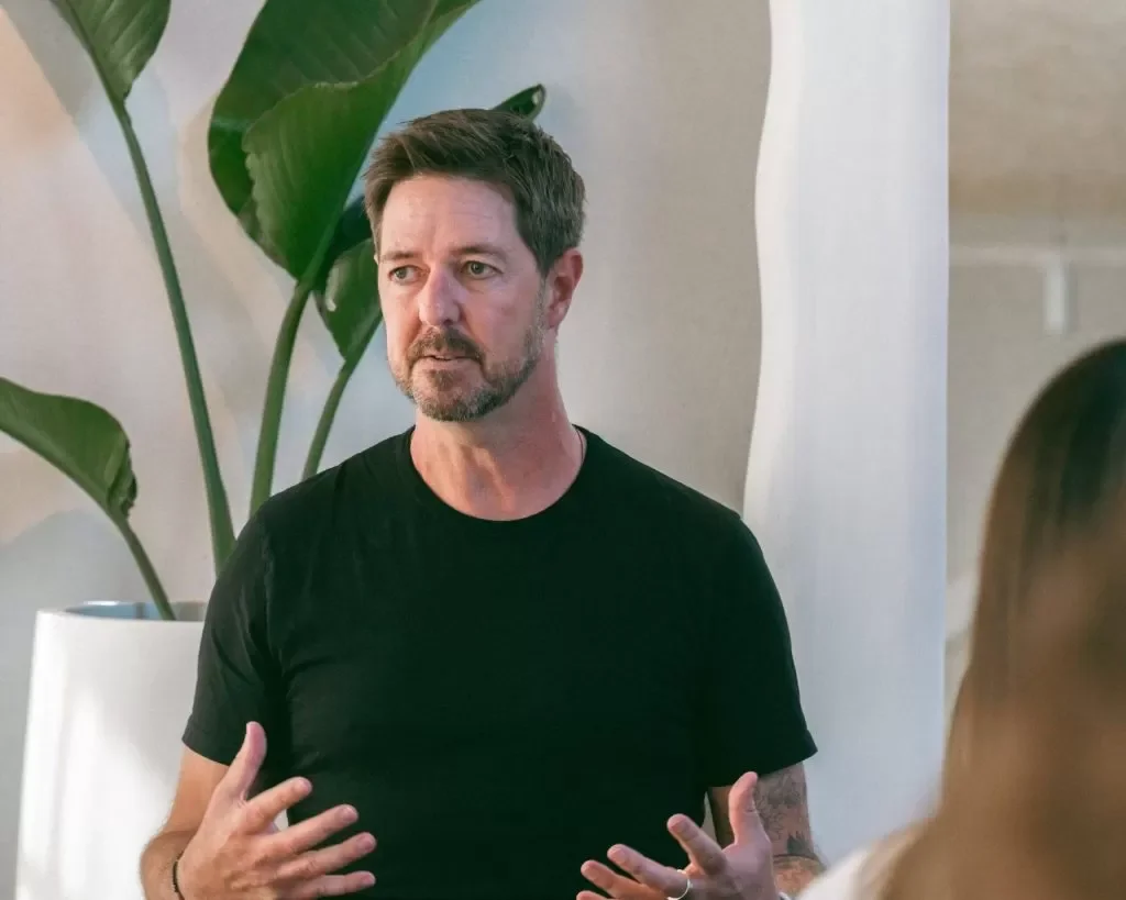 A man with short brown hair and a beard, wearing a black t-shirt, is speaking or explaining something in front of a large leafy plant and a white and beige background.