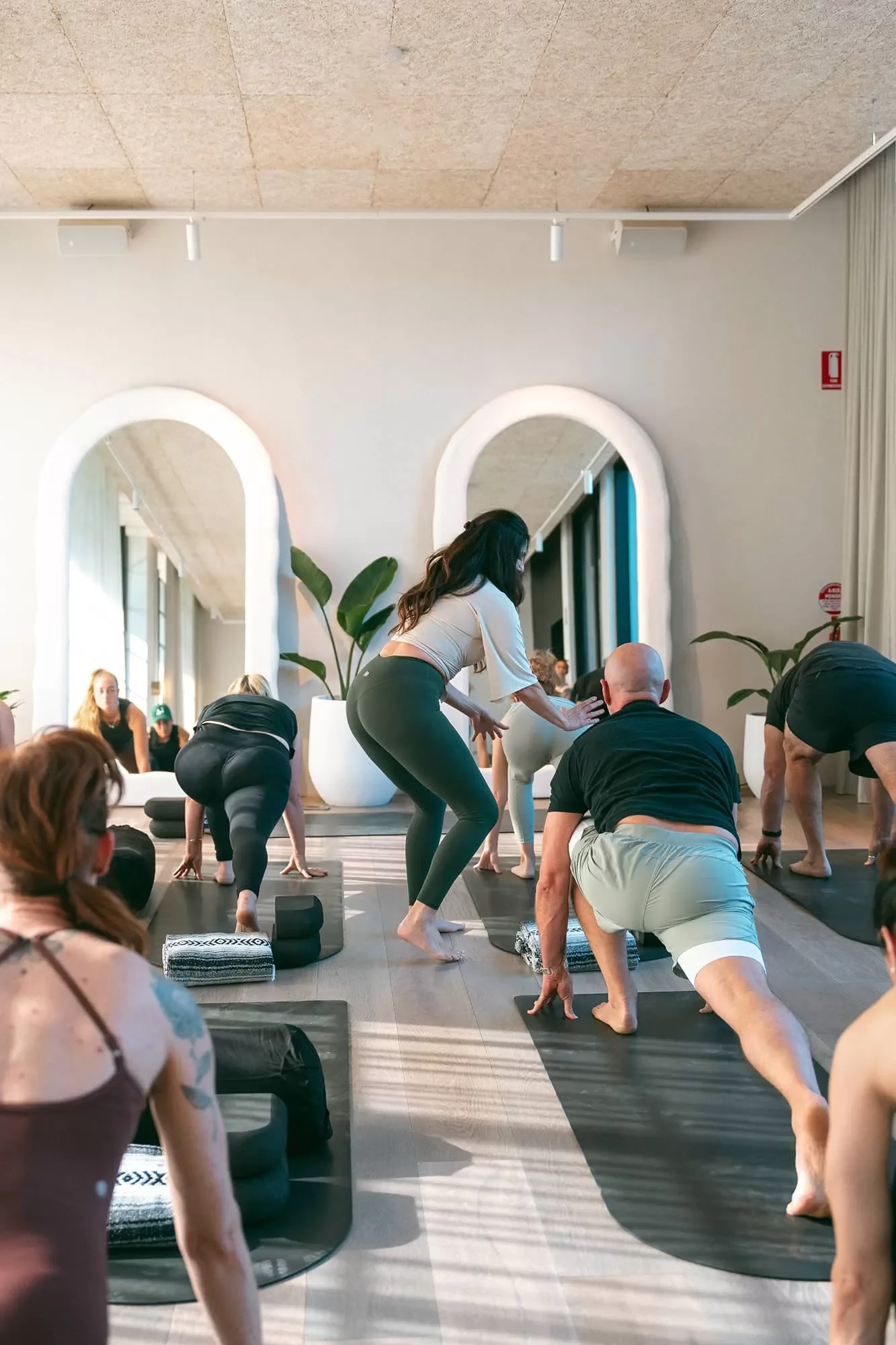 People participating in a yoga class in a bright, modern studio with large mirrors, potted plants, and a light-colored wood floor.