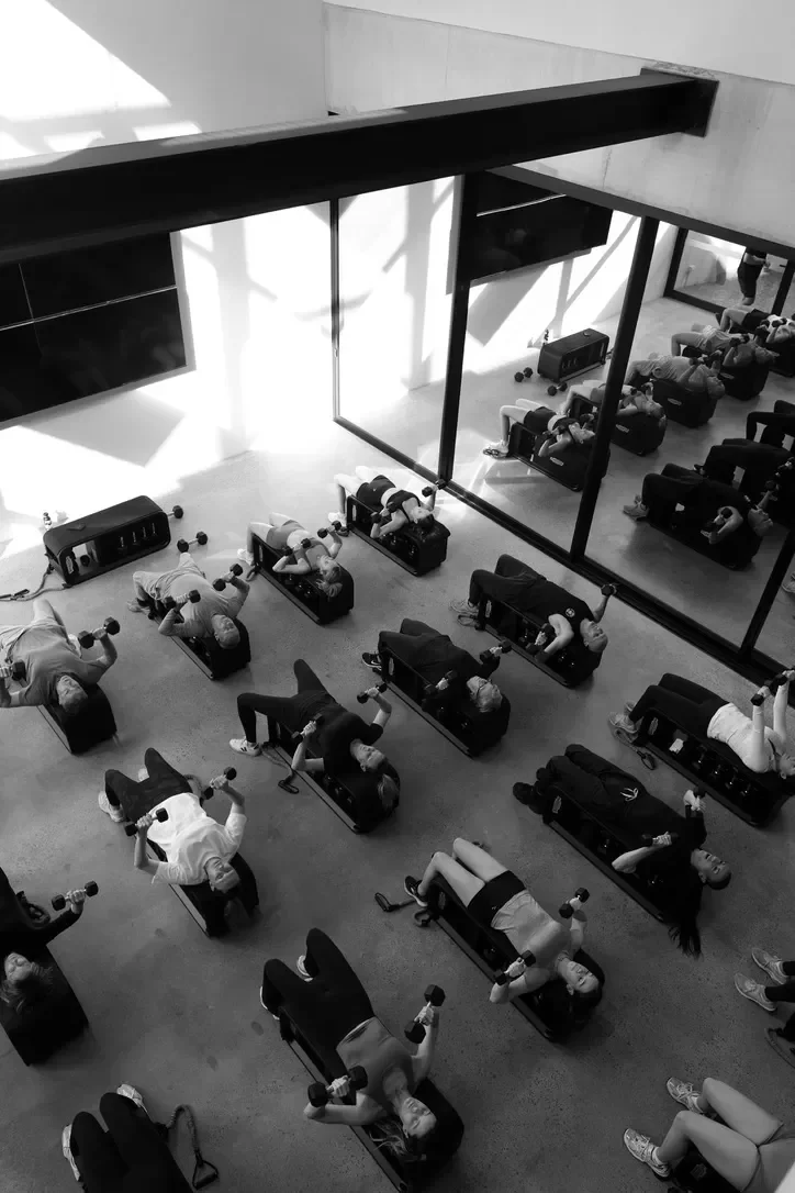 Group fitness class with people lying on workout benches, lifting dumbbells, in a gym with large mirrors and a bright interior